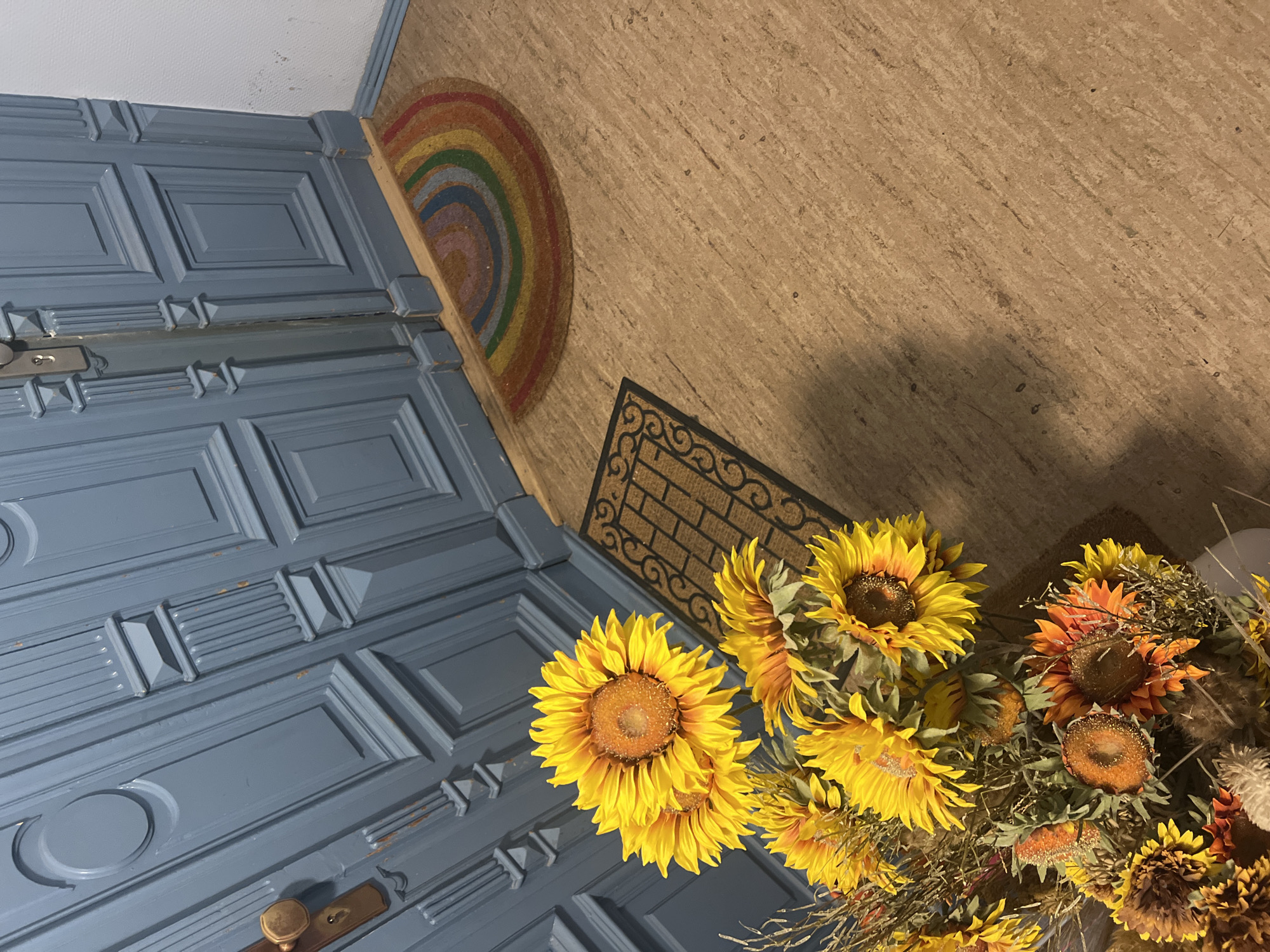 A close-up, high-angle shot shows a bouquet of sunflowers and dried fall foliage in a pot, placed on a light brown, textured floor. To the left, a large, blue, paneled door with ornate gold hardware is visible. In the bottom right corner of the door, a semi-circular doormat with rainbow-colored stripes is partially visible. Next to the rainbow mat is a rectangular doormat with a decorative pattern. The scene appears to be an entryway, possibly in a residential building in Berlin, Germany, given the architectural style of the door. No people are present in the image.