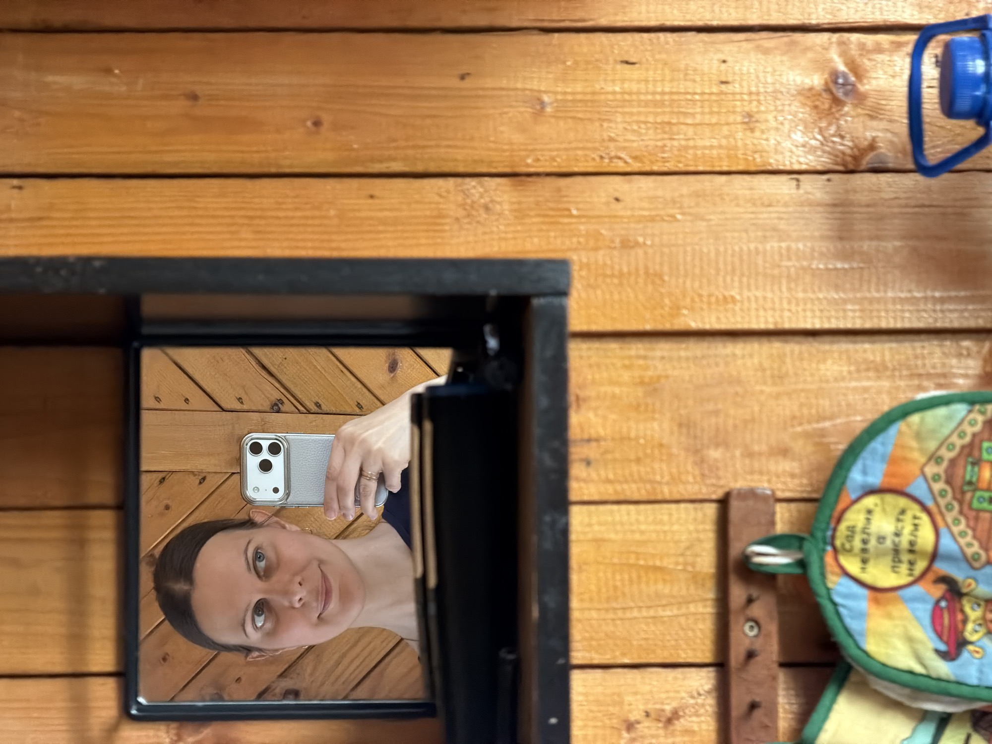 The image presents a medium shot of a room with wood-paneled walls. In the lower left, a black-framed mirror reflects a woman holding a cell phone. Her face is visible, and she appears to be smiling slightly. The phone she holds is a modern smartphone with a triple-lens camera system. To the right of the mirror, a blue bottle cap with a handle is affixed to the wall. Below that is a wooden peg rack and a round pot holder. The style of the interior suggests a rustic, possibly rural, setting. There is no explicit information present in the image to suggest Izhevsk, Russia or any other location.