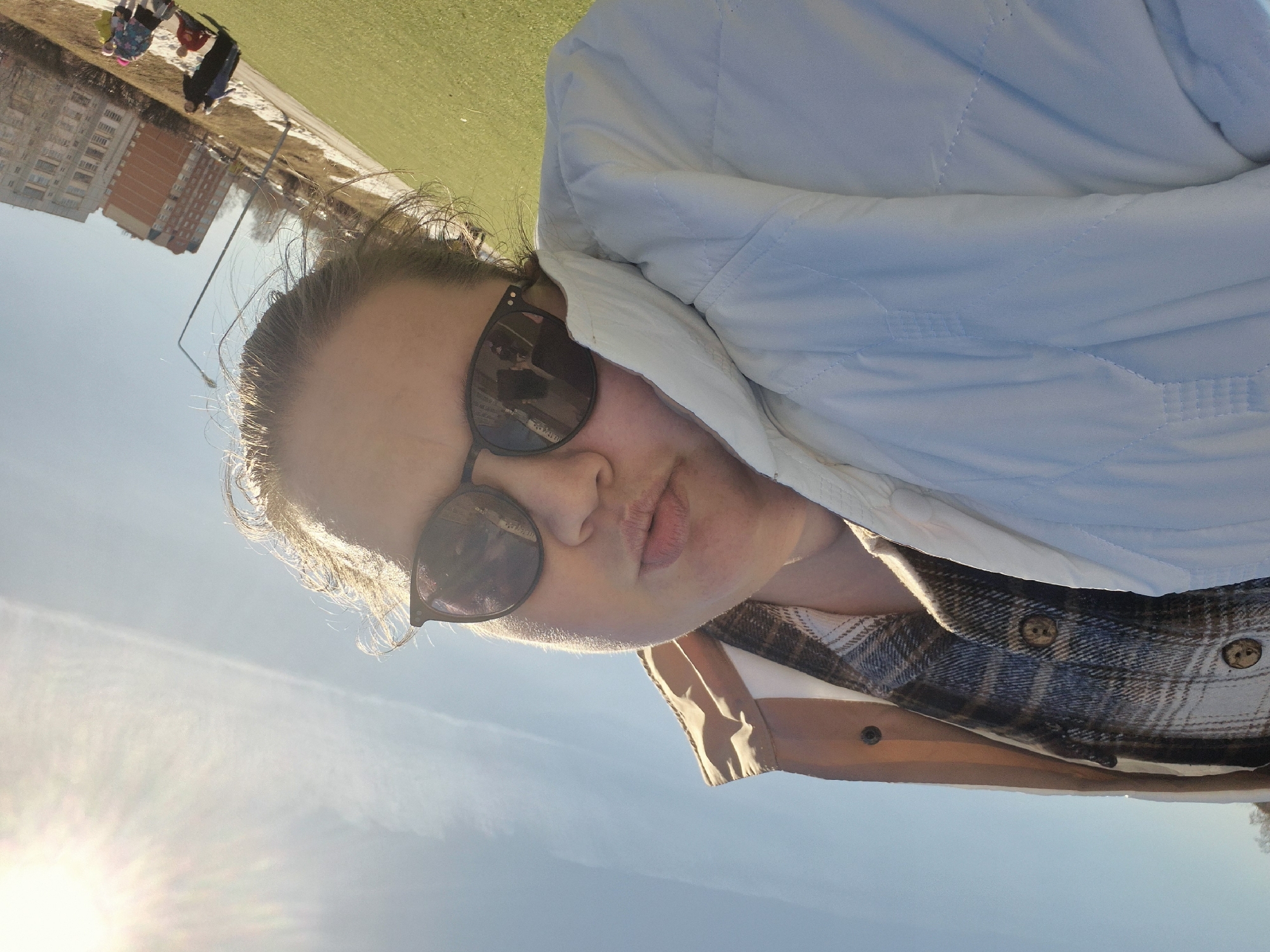 A woman wearing sunglasses and a white jacket is taking a selfie outdoors on a sunny day. She is also wearing a plaid shirt. The background shows a cityscape with apartment buildings, a grassy field, and other people in the distance. The specific location is Chaikovsky, Russia.