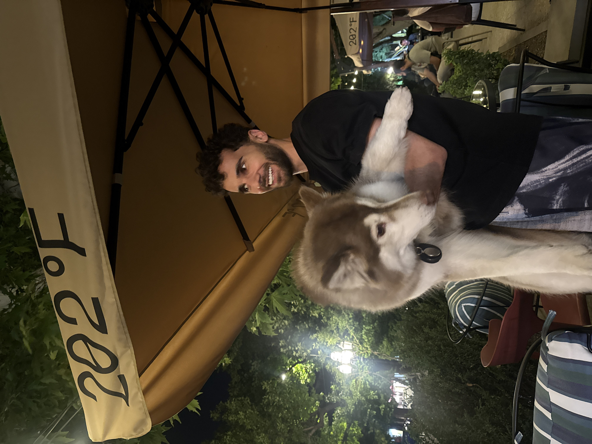 A man with curly dark hair and a beard is holding a husky dog in his arms. The man is wearing a black t-shirt and patterned pants. The dog has brown and white fur. They are standing outdoors at night, under a tan umbrella with "202°F" written on it. In the background, there are trees with lights, other people sitting at tables, and more umbrellas. The scene appears to be an outdoor seating area of a restaurant or cafe. The umbrella with the temperature marking suggests this is likely located in Yerevan, Armenia, as it is a common feature of outdoor cafes and restaurants there.