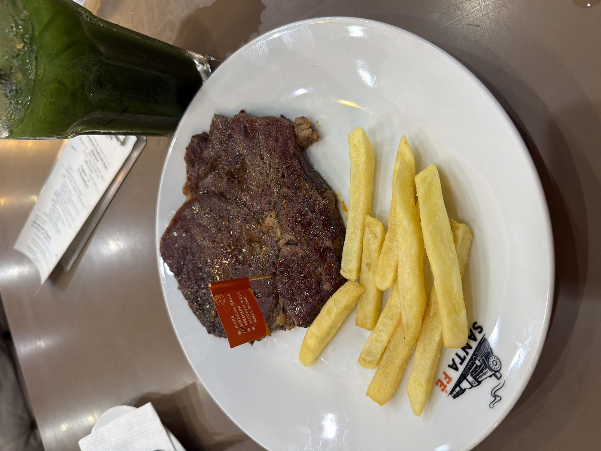 A plate of steak and fries, along with a green beverage, sits on a table. The plate is white and displays the "Santa Fe" logo. A small red flag is stuck into the steak, indicating its level of doneness. The location is Baan Chaweng, Thailand. There are no people visible in the image.