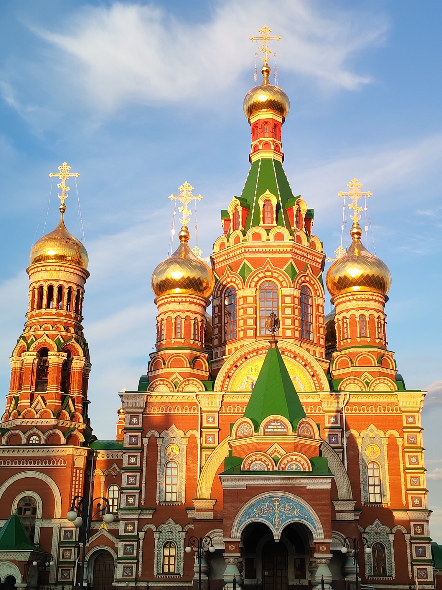 The image depicts a close-up view of the Church of the Archangel Gabriel in Yoshkar-Ola, Russia. The architecture is characterized by its ornate design, featuring multiple golden onion domes topped with Orthodox crosses. The facade is constructed primarily of red brick, accented with decorative white and green elements, including arched windows and intricate mosaic patterns. The structure is illuminated by the warm, golden light of what appears to be sunset or sunrise, casting long shadows and highlighting the architectural details. The sky above is a clear blue with scattered white clouds. No people or animals are visible in the frame. The scene is static and focuses solely on the exterior of the religious building.
