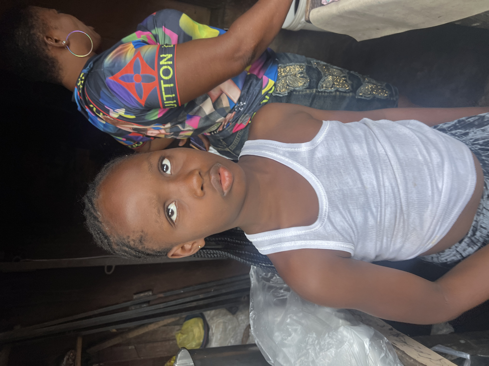 A portrait-oriented image captures two Black individuals, a young girl and an adult woman, in an informal indoor or semi-sheltered setting, likely in Nkpor, Nigeria.
In the foreground and center, a young girl with braided hair looks directly at the camera with a neutral yet attentive expression. She is wearing a simple white ribbed tank top and patterned shorts or a skirt, partially visible. She has small stud earrings in her ears.
Behind the girl and slightly to her left, an adult woman is partially visible. She wears a striking, multi-colored short-sleeved top featuring patterns reminiscent of the Louis Vuitton monogram, with the word "VUITTON" clearly visible in yellow lettering on a black band on her right sleeve. Her attire also includes dark-colored bottoms, possibly jeans, with decorative gold-colored embellishments. A large, iridescent hoop earring is prominent on her left ear. The woman is turned slightly away from the camera, gazing towards her right, suggesting a candid moment.
The background is somewhat dark and indistinct, showing elements such as what appears to be a wall or shelving structure, a light-colored cushion or furniture piece behind the woman, and various objects on the ground including a clear plastic bag and another bag containing a yellow item. The lighting suggests it is daytime. The scene conveys a casual, everyday moment.
