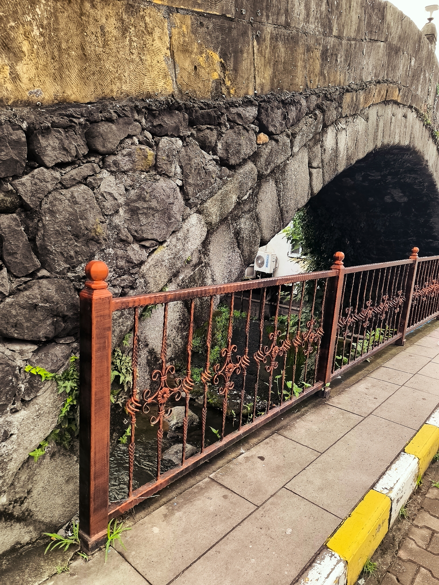 The image depicts an arched stone bridge in Makho, Georgia. The bridge's upper section is constructed from large, weathered stones with a golden-yellow hue, possibly sandstone. Below this, the arch and the retaining wall are built from rough-hewn, dark grey stones. A reddish-brown metal railing with decorative scrollwork runs along the walkway beside the bridge. The walkway itself is paved with rectangular concrete tiles, bordered by a curb painted with alternating yellow and white stripes. A small stream flows beneath the bridge, with green vegetation growing along its banks and on the bridge's stone structures. An air conditioning unit is visible on a white building peeking out from under the arch. No people are present in the image. The scene is an outdoor view of a section of the bridge and its surrounding elements.
