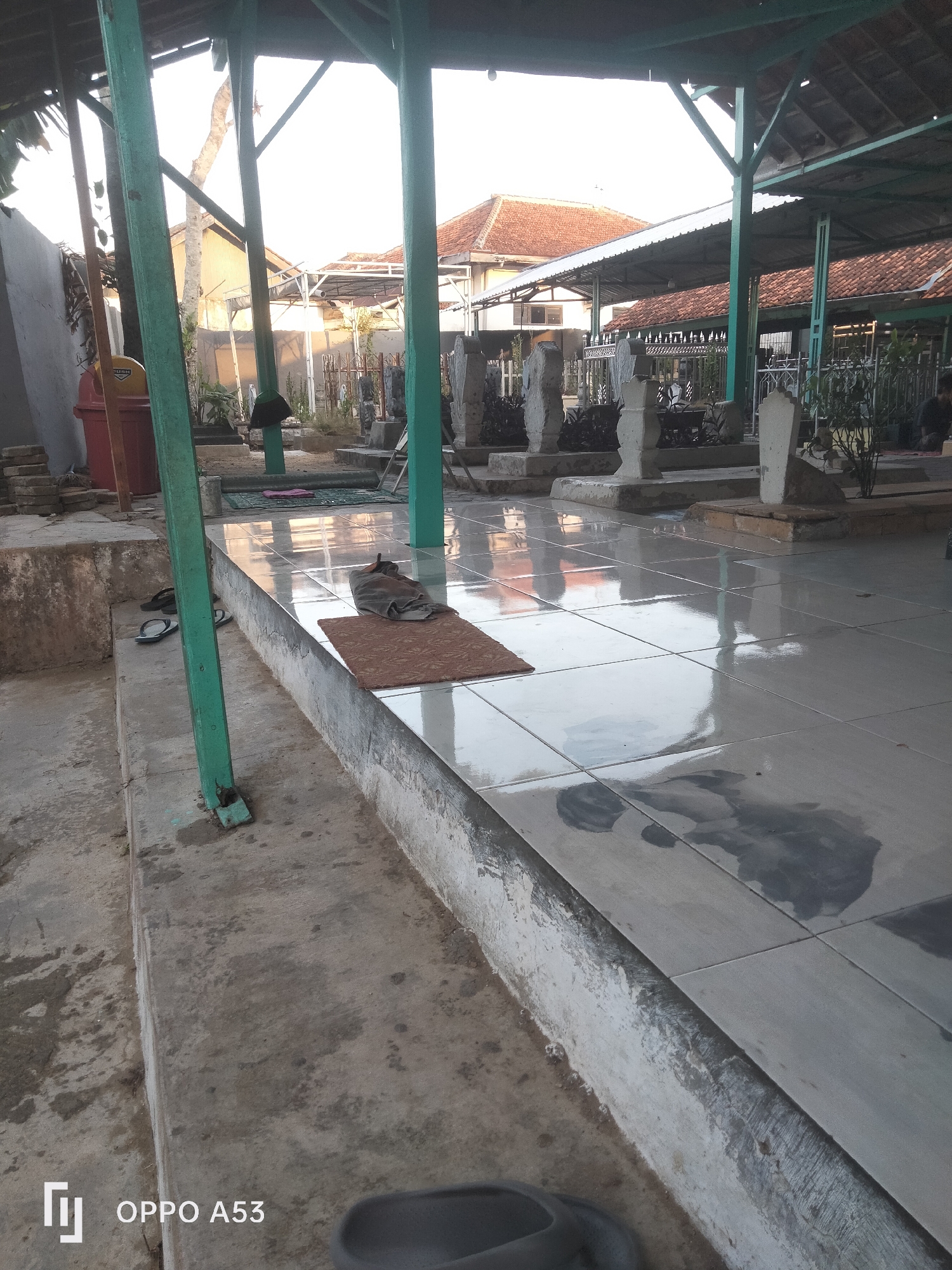 A medium shot captures an outdoor sheltered area, likely a cemetery or shrine, with a tiled floor that reflects light and water. In the foreground, a grey mat is placed on the tiled surface, with a dark cloth or towel lying on top. To the left, a concrete embankment is visible, with several pairs of black flip-flops scattered on it. Green metal posts support the shelter's roof, which appears to be made of corrugated metal and tiles. Beyond the sheltered area, several gravestones are arranged, some with vegetation growing around them. In the background, buildings with red tile roofs and a tall, slender tree are visible. A red trash bin is also present to the left. The overall scene suggests a quiet, potentially humid environment, common in tropical locations like Badung, Indonesia, due to the visible water on the tiles and the vegetation.