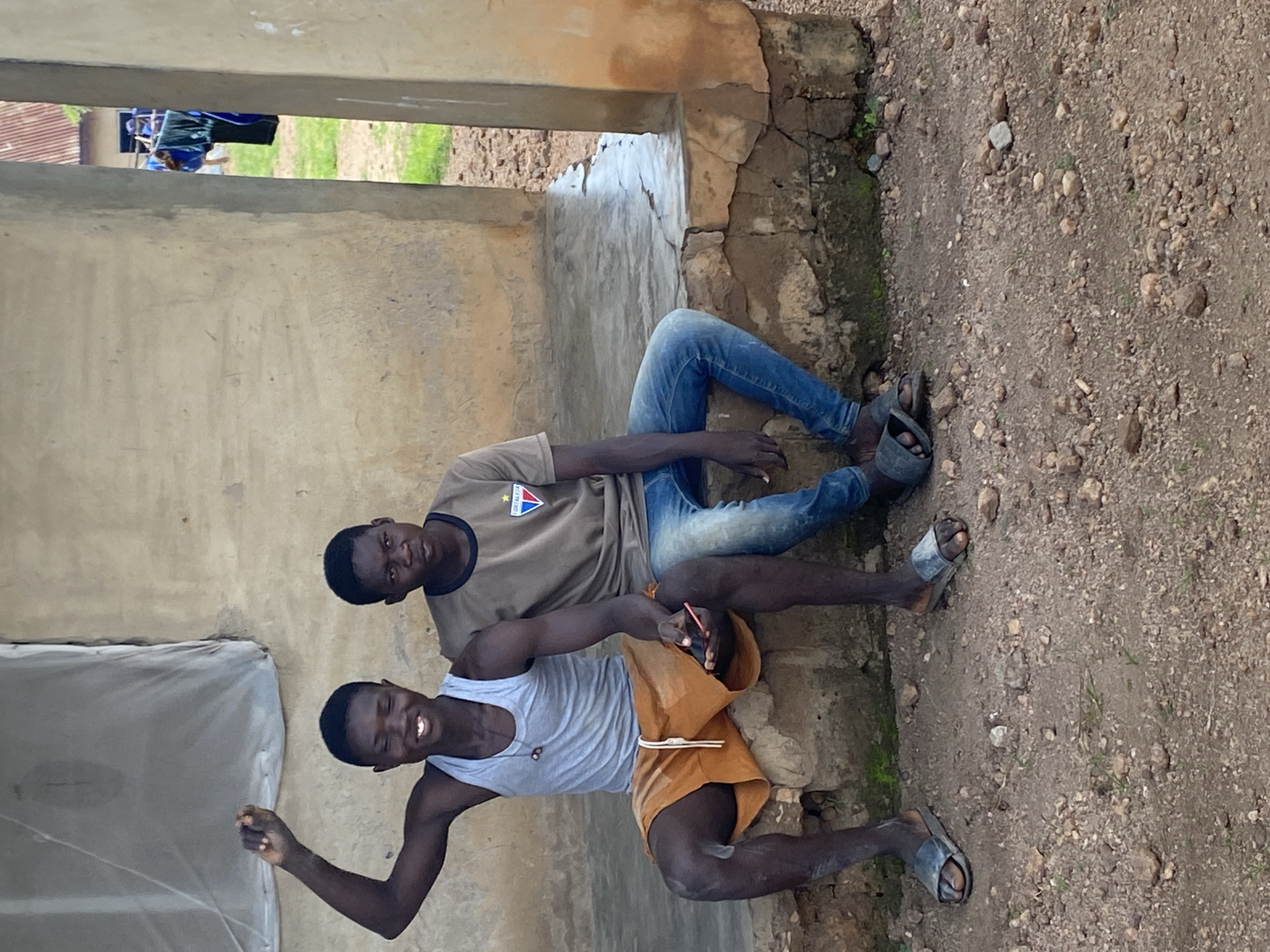 Two young Black males are sitting on a concrete ledge outside a rough-textured, beige building. The male on the left, wearing a grey tank top and orange shorts, is smiling and has his left arm raised. The male on the right, wearing a brown t-shirt and blue jeans, is looking forward. Both are wearing sandals. The ground in front of them is covered in dirt and small rocks. Behind them, clothes are hanging on a line. The image appears to have been taken outdoors during the day.