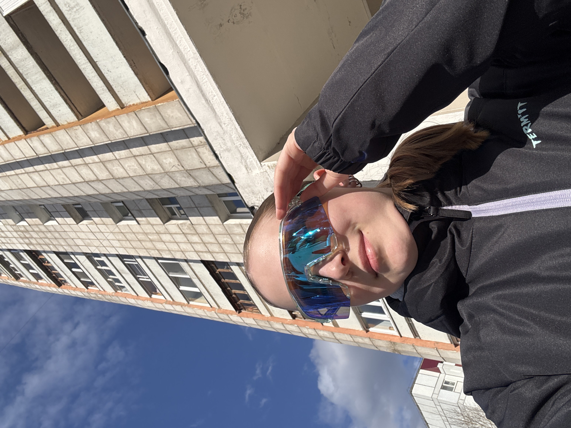 This outdoor portrait, taken in Chaikovsky, Russia, features a young person on a sunny day. The subject, viewed from the upper chest, is wearing a black jacket with a light-colored zipper and collar. Visible white text on the jacket's sleeve reads "TERMIT." They sport large, wrap-around, reflective sunglasses that show iridescent blue and orange/yellow hues, reflecting the sky and surroundings. One hand is raised, appearing to adjust the sunglasses on their forehead. The visible part of their face, from the mouth downwards, shows a neutral or relaxed expression.
In the background, a towering, light-colored concrete or stucco building dominates the scene, featuring multiple floors with repetitive window and balcony structures, indicative of residential or institutional architecture. Part of a smaller, white building is also visible in the lower right. The sky above is a clear, bright blue with scattered white clouds, suggesting fine weather and bright daylight. The image is tilted, giving a dynamic, upward-looking perspective that emphasizes the height of the main building.