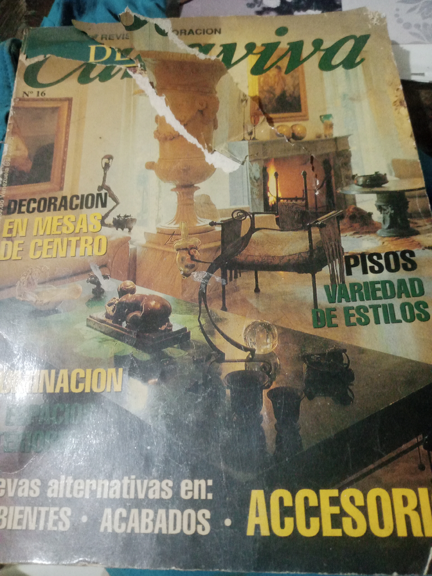 The image shows the cover of a magazine titled "DECORACION VIVA" with issue number 16. The magazine has visible wear and tear, including rips and creases. The cover features a photograph of interior decoration elements. In the foreground, there are several decorative objects, including a bronze sculpture of a dog and a globe. In the mid-ground, a fireplace with a fire burning is visible, along with a chair and a table. The text on the cover advertises "DECORACION EN MESAS DE CENTRO" (decoration for coffee tables), "PISOS VARIEDAD DE ESTILOS" (floors variety of styles), and "ACCESORIOS" (accessories). The magazine's origin is indicated as Venezuela. No specific location in Santa Ana, Venezuela, can be identified from the image.