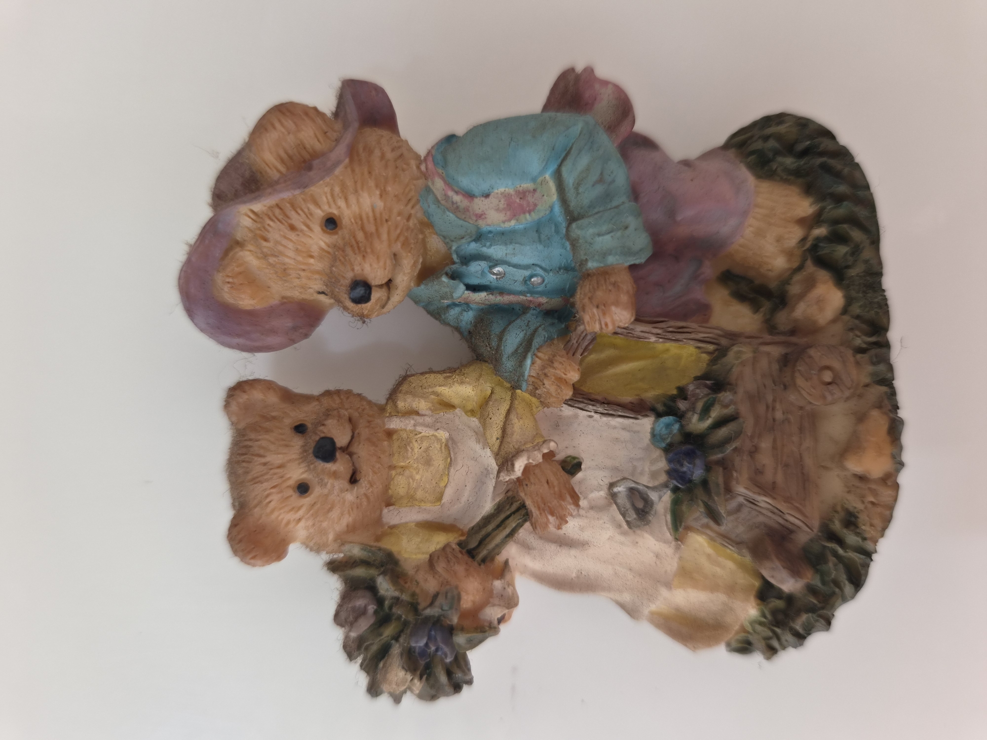A figurine of two teddy bears stands against a white background. The bear on the left wears a yellow dress and a white apron, and holds a bouquet of flowers. The bear on the right wears a blue shirt and a purple hat. The bears are standing on a base decorated with grass and a small wooden cart.