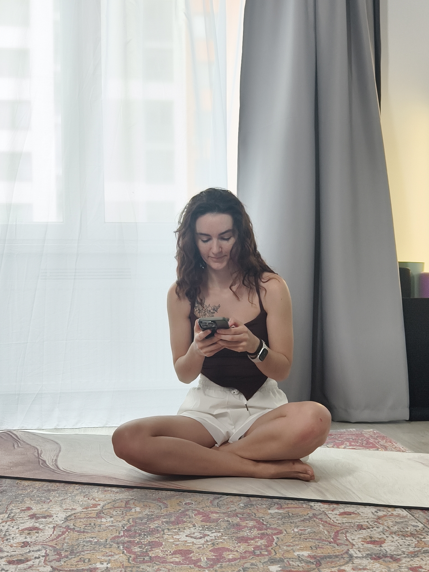 A young woman is seated cross-legged on a yoga mat, looking down and interacting with a smartphone. She is wearing a dark brown tank top and white shorts. She has long, curly brown hair and a tattoo on her chest. The background consists of white curtains to her left and gray curtains to her right. A small stack of yoga mats of different colors can be seen in the background to her right. The scene is set indoors, likely in her apartment, judging by the furnishings and personal items. The floor beneath the yoga mat is covered by a patterned rug. The location is Tyumen, Russia.