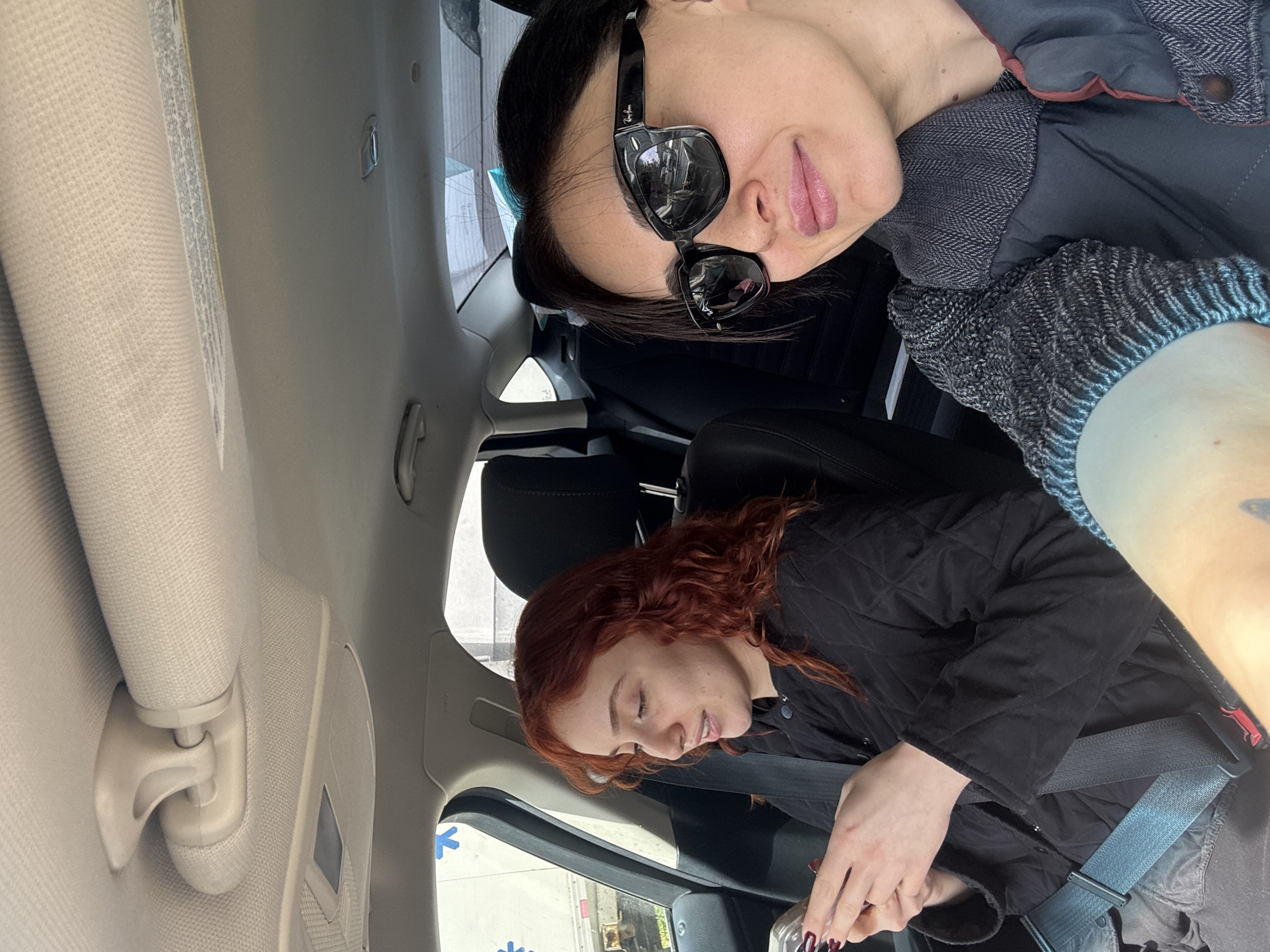 This media file features two individuals inside a car, photographed from a perspective looking from the rear passenger area towards the front. The image is rotated 90 degrees clockwise.
In the foreground (top of the rotated image), an adult female with dark hair is visible, wearing black Ray-Ban sunglasses and a dark jacket with a textured grey collar and knitted cuffs. She has pink lipstick and appears to be resting or looking towards the side.
In the background (bottom of the rotated image), a younger female with red, wavy hair is seated, wearing a black quilted jacket and a seatbelt. She is looking downwards, holding a cup or bottle with her hands, which have dark nail polish.
The interior of the car is primarily beige and light grey, showing the headliner, sun visor, and seat elements. Through the window beside the red-haired person, a glimpse of the outside world is visible, including what appears to be part of a building or a white vehicle, and several blue arrow symbols on an adjacent surface. The setting suggests daytime travel or a stop within a vehicle. Both individuals maintain a relaxed or neutral expression. The location context provided is Tbilisi, Georgia.