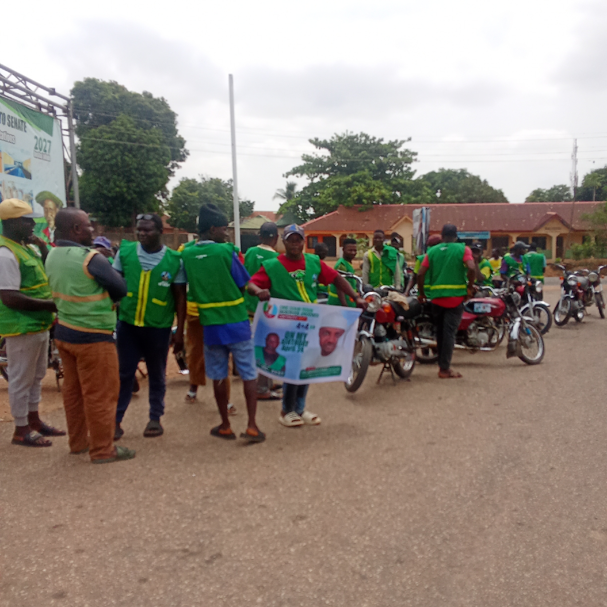 A group of people wearing green and yellow vests are gathered on a paved road in Akwanga, Nigeria. Some are standing while others are near parked motorcycles. One person holds a poster featuring two individuals and the text "ON MY MANDATE APRIL 24". In the background are trees and a building with a red roof. A banner with the words "TO SENATE" and "2027" is partially visible on the left. The sky is overcast.