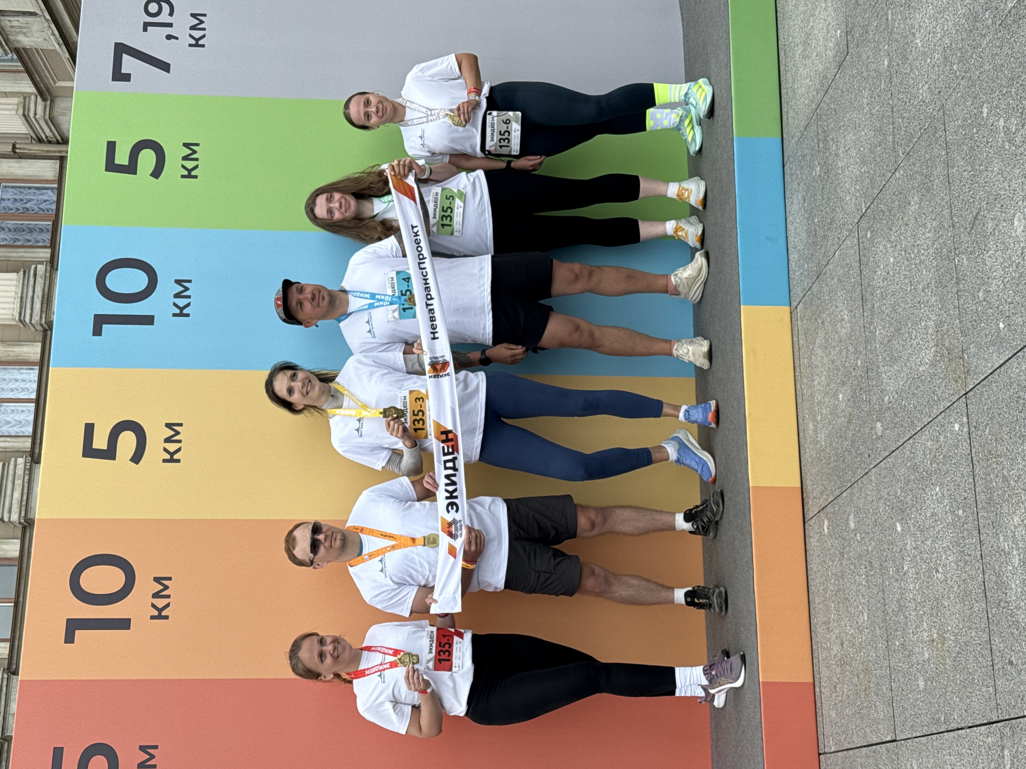 Six individuals, four women and two men, are posing for a photograph on a raised platform in front of a colorful backdrop displaying various kilometer distances associated with a race. The backdrop features panels in orange, yellow, light blue, green, and grey, with large black numbers indicating "10 KM", "5 KM", and "7.19 KM". The individuals are all wearing white t-shirts, bib numbers, and medals, suggesting they have participated in a running event.
From left to right:
The first woman on the left is wearing black leggings and white socks, holding a medal. Her bib number is "135-1".
The first man is wearing dark shorts, sunglasses, and a cap, holding a medal.
The second woman is holding a white banner with text that appears to be in Russian. She is wearing dark leggings and a medal.
The second man is wearing dark shorts and a medal.
The third woman is wearing dark leggings and a medal. Her bib number is "135-5".
The fourth woman is wearing dark leggings and a medal. Her bib number is "135-6".
The banner held by the second woman reads "ЭКИДЕН" and "НеваТрансПроект", which translates to "EKIDEN" and "NevaTransProject". The backdrop's color scheme and the presence of race distances suggest this is an outdoor event. The building visible in the background suggests an urban setting, possibly in Saint Petersburg, Russia, given the likely sponsor "NevaTransProject" and the commonality of such running events in large Russian cities. The ground surface in front of the platform is paved with grey granite tiles. The overall scene is brightly lit, indicating daytime.