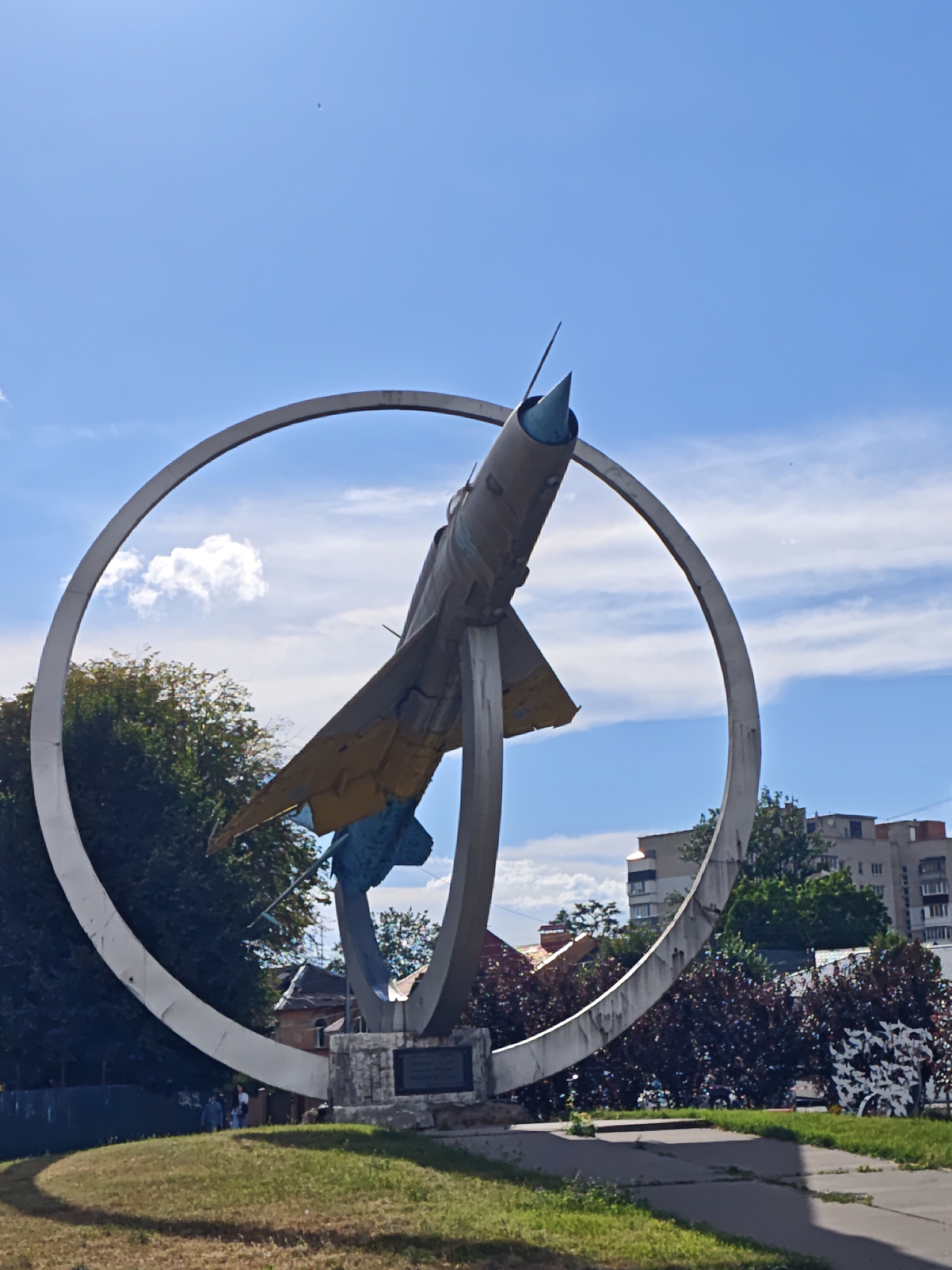 The image shows a monument to an aircraft, likely a MiG-21, on a pedestal with a large metal ring behind it. The monument is located in a park-like area with green grass, trees, and some buildings in the background. The sky is blue with scattered clouds. Several people are visible in the background, standing near the monument. The monument appears to be in a public space, possibly a memorial or a historical landmark. The aircraft is tilted upwards, as if in flight, with its nose cone painted blue. The metal ring is silver and is positioned behind the aircraft. The pedestal is made of stone and has a plaque on it. The overall scene suggests a sunny day with good visibility. The monument is a prominent feature in the image, drawing the viewer's attention due to its size and unique design. The presence of people indicates that it is accessible to the public and likely a place of interest for visitors. The surrounding environment, with its greenery and sky, creates a serene and pleasant atmosphere. The buildings in the background suggest an urban setting, indicating that the monument is situated within a city. The monument's design, featuring an aircraft within a ring, could symbolize flight, achievement, or remembrance. The blue nose cone of the aircraft might have symbolic meaning, perhaps representing the sky or a specific aspect of aviation. The presence of a plaque on the pedestal further suggests that the monument has a historical or commemorative purpose.