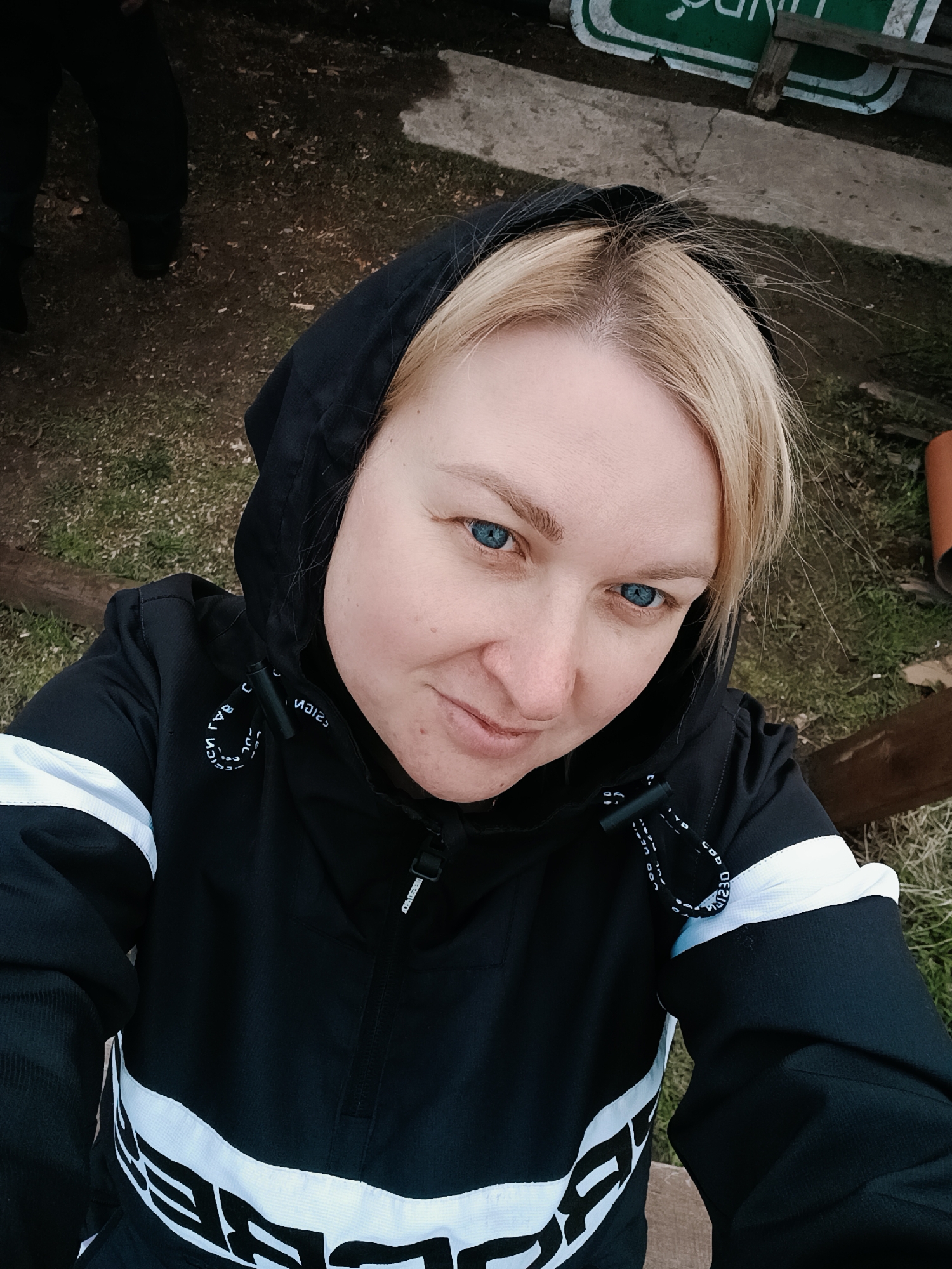 A woman with blonde hair and striking blue eyes takes a selfie in an outdoor setting, presumably in Chaikovsky, Russia. She looks directly at the camera with a calm, subtle smile. She is wearing a black hooded jacket featuring distinct white horizontal stripes and white text that prominently displays "DEFEND" across the front. Additional white text, appearing to be "DEFEND" reversed or similar wording, is visible on the black and white drawstrings of her hood, and a small tag on the jacket's zipper pull also has text.
The background consists of a mix of dry grass and dirt, with patches of concrete. In the upper left, the lower legs and feet of another person, clad in dark clothing and shoes, are partially visible. A wooden log or bench is situated to the right of the woman. Above her head, a partially obscured, upside-down green sign with white text is present, alongside an orange pipe-like object on the ground. The lighting is diffused, suggesting an overcast day.