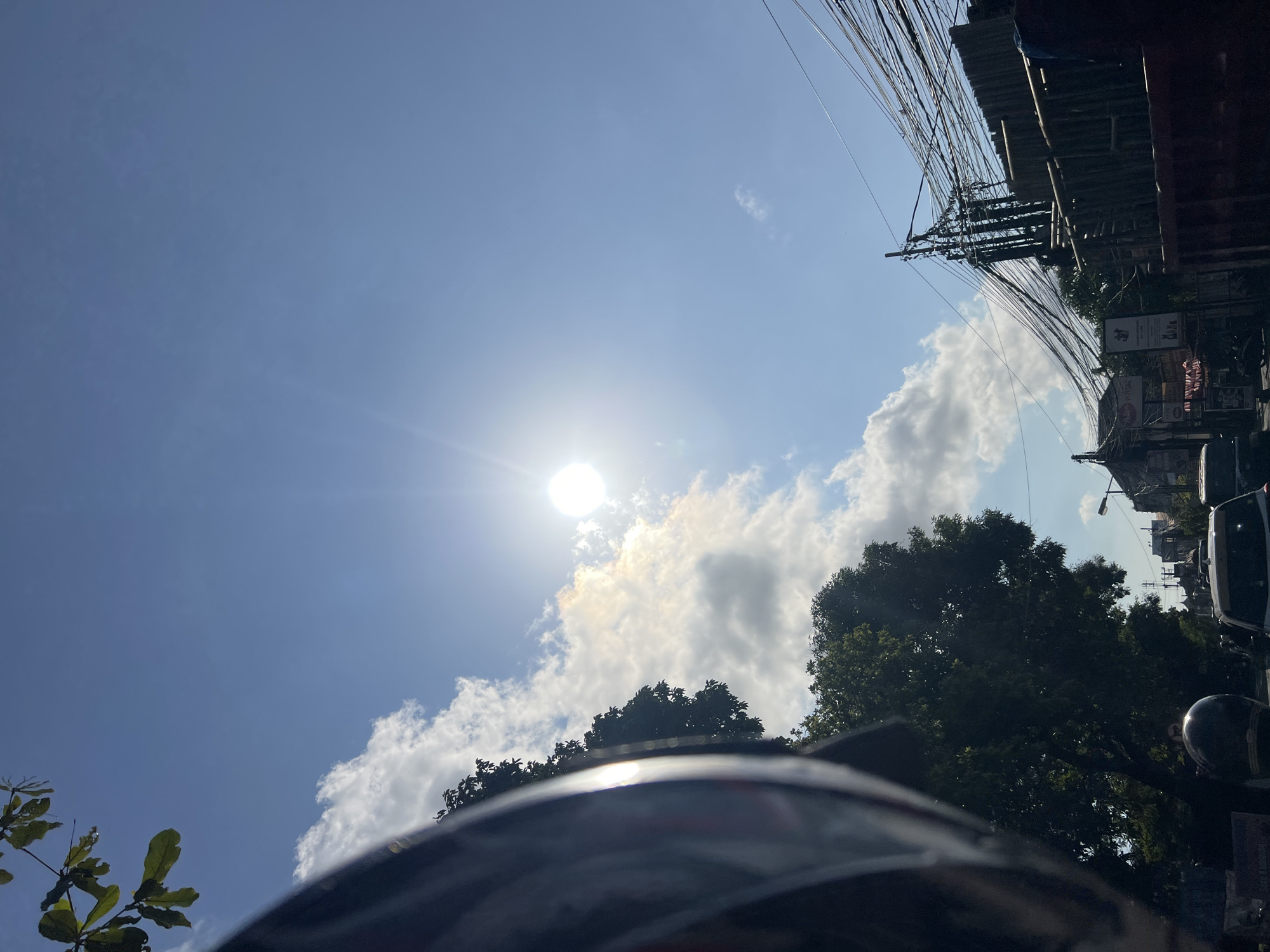 The image captures a bright, sunny day in Denpasar, Indonesia, likely a street scene. The sun is high in the sky, casting a bright glare and creating lens flare. Scattered white clouds drift across the clear blue sky. In the foreground, the side of a dark helmet and a portion of a motorcycle's handlebars are visible, suggesting the photo was taken from the perspective of a rider. Lush green trees line the street, providing shade. Overhead, a dense network of electrical wires crisscrosses between poles, a common sight in many Indonesian cities. Buildings are visible in the background, some appearing to be shops with signage, though the text is not clearly legible. The street below appears to be busy with traffic, with the tops of vehicles, including cars and possibly another motorcycle, visible. The overall impression is a snapshot of daily life in a bustling urban environment under a strong tropical sun.
