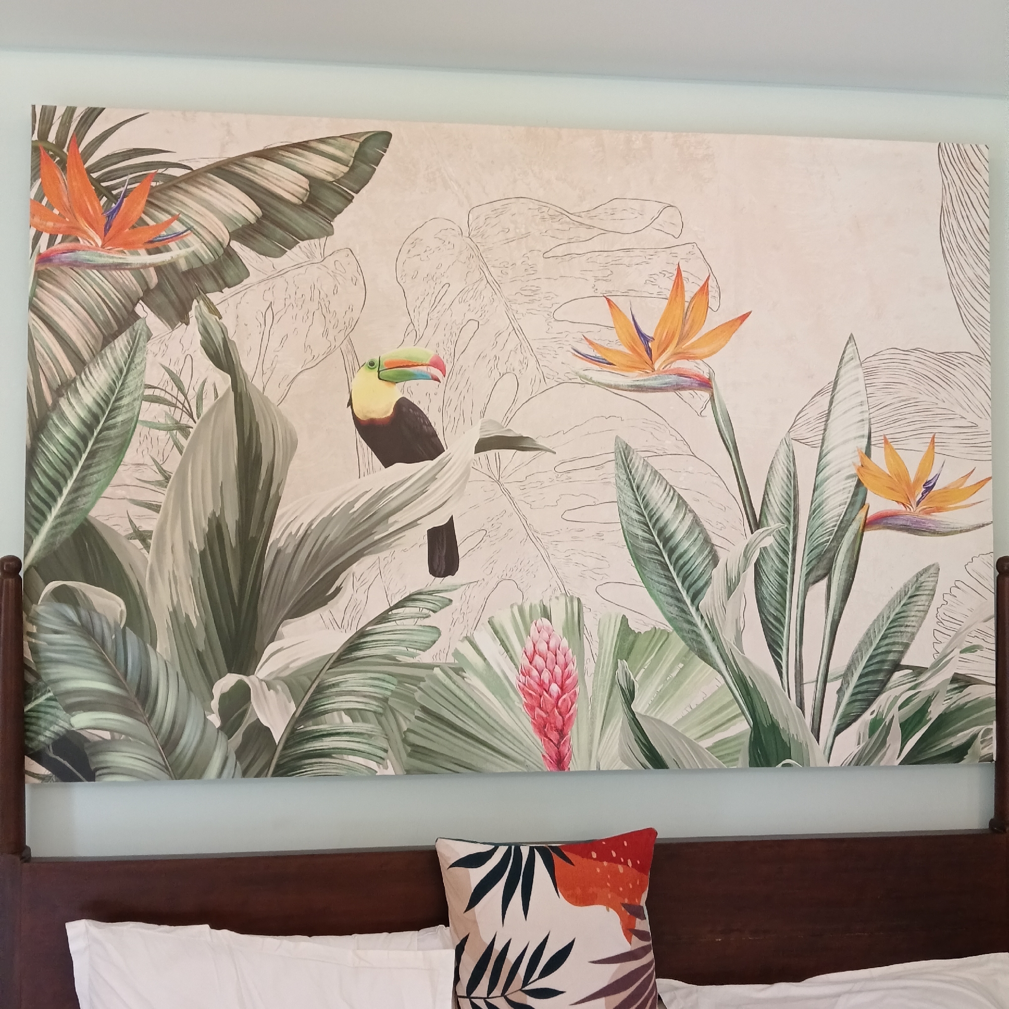 The image depicts a section of a bedroom interior. A dark wooden headboard sits above two white pillows and a decorative cushion. The cushion has a light-colored background with botanical patterns in black, orange, and maroon hues. Above the headboard is a large framed print with a tropical motif. The print showcases illustrated images of large leaves, bird-of-paradise flowers in orange and yellow, a pink ginger flower, and a toucan. Some of the leaves in the print are fully colored, while others are represented in outline form. The background wall is painted a light blue. The location is specified as Calangute, India.