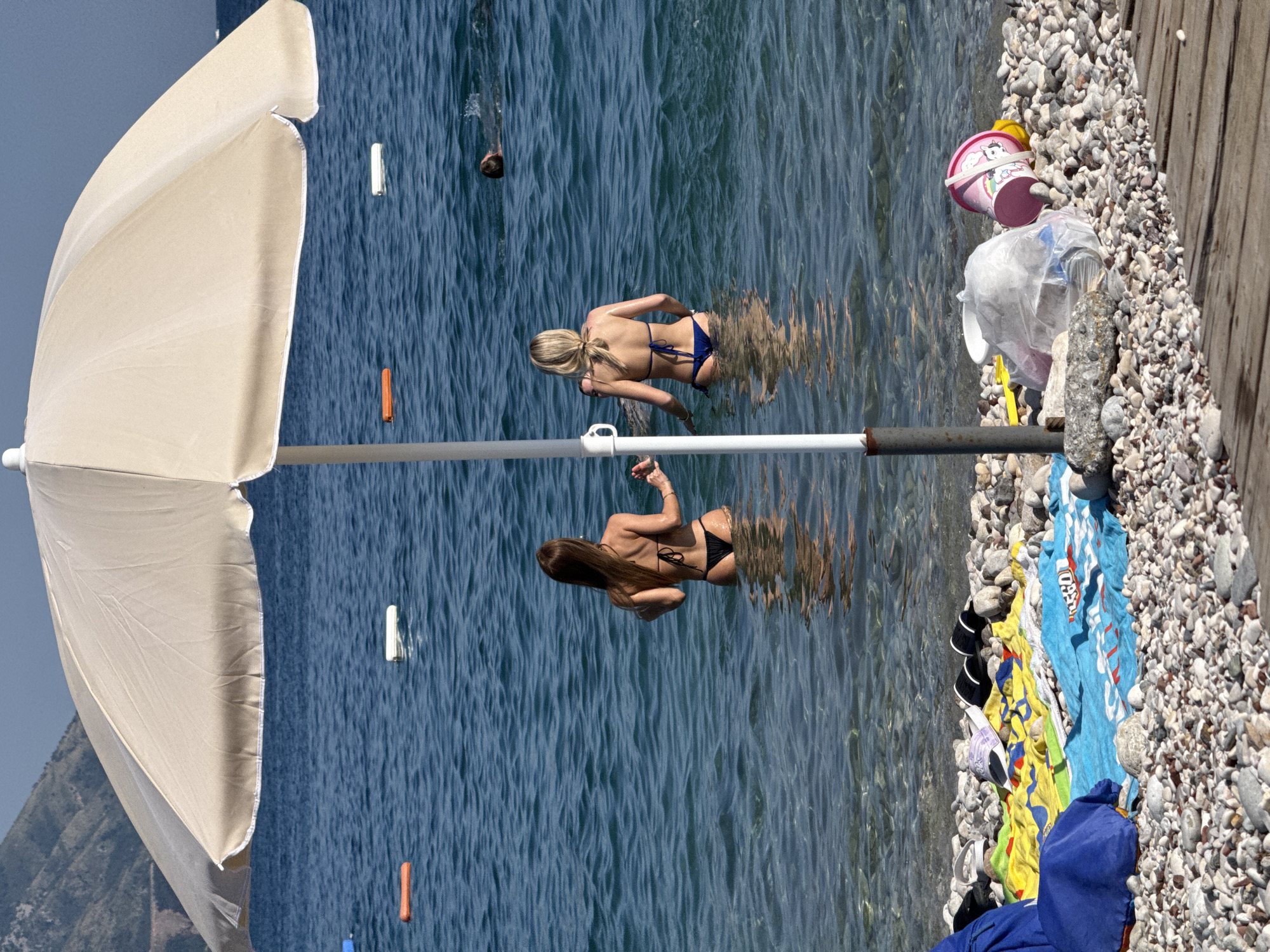 The image depicts a sunny day at a pebble beach in Budva, Montenegro. Two women are in the shallow, clear blue water. One woman, with blonde hair tied back, wears a blue bikini and stands facing away from the camera, her back to the viewer. The other woman, with long brown hair, also wears a black bikini and stands facing away, her arms slightly raised. Both women are partially submerged in the water, which reflects the light and creates a rippled effect.
To the right of the water, a wooden walkway borders the pebble beach. Scattered on the pebbles are various items, including colorful towels, a pink child's bucket with a cartoon character, and what appears to be a plastic bag. A large, cream-colored beach umbrella is prominently positioned on the left side of the frame, casting a shadow. In the background, the blue sky meets a distant, mountainous landscape, suggesting a coastal environment. The scene appears to be a typical summer day at the beach, with an emphasis on leisure and relaxation. The presence of beach items and the clear water indicate warm, pleasant weather suitable for swimming and sunbathing.