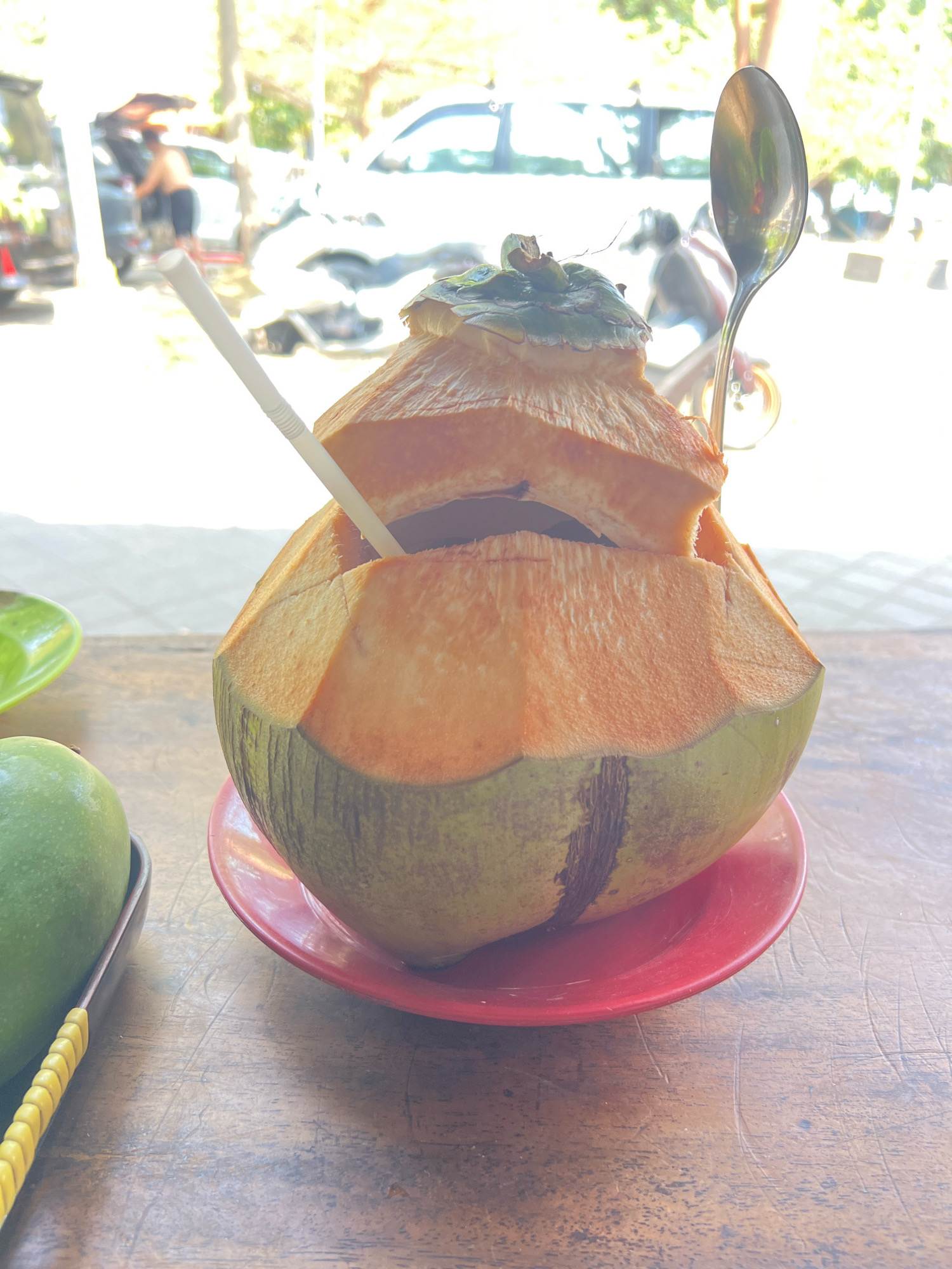 A close-up, eye-level shot shows a halved coconut on a red plate, resting on a wooden table. A white straw is inserted into the coconut, and a spoon stands upright behind it. To the left, a green fruit, likely a mango, is partially visible on a brown tray with a yellow handle. In the blurred background, a person is seen near an open car trunk, with other vehicles and foliage suggesting an outdoor setting. The lighting is bright and appears to be natural sunlight, creating a washed-out effect on the background details. The scene suggests a casual refreshment break, possibly at a beachside cafe or stall in Kuta, Indonesia, given the tropical fruit and outdoor ambiance.