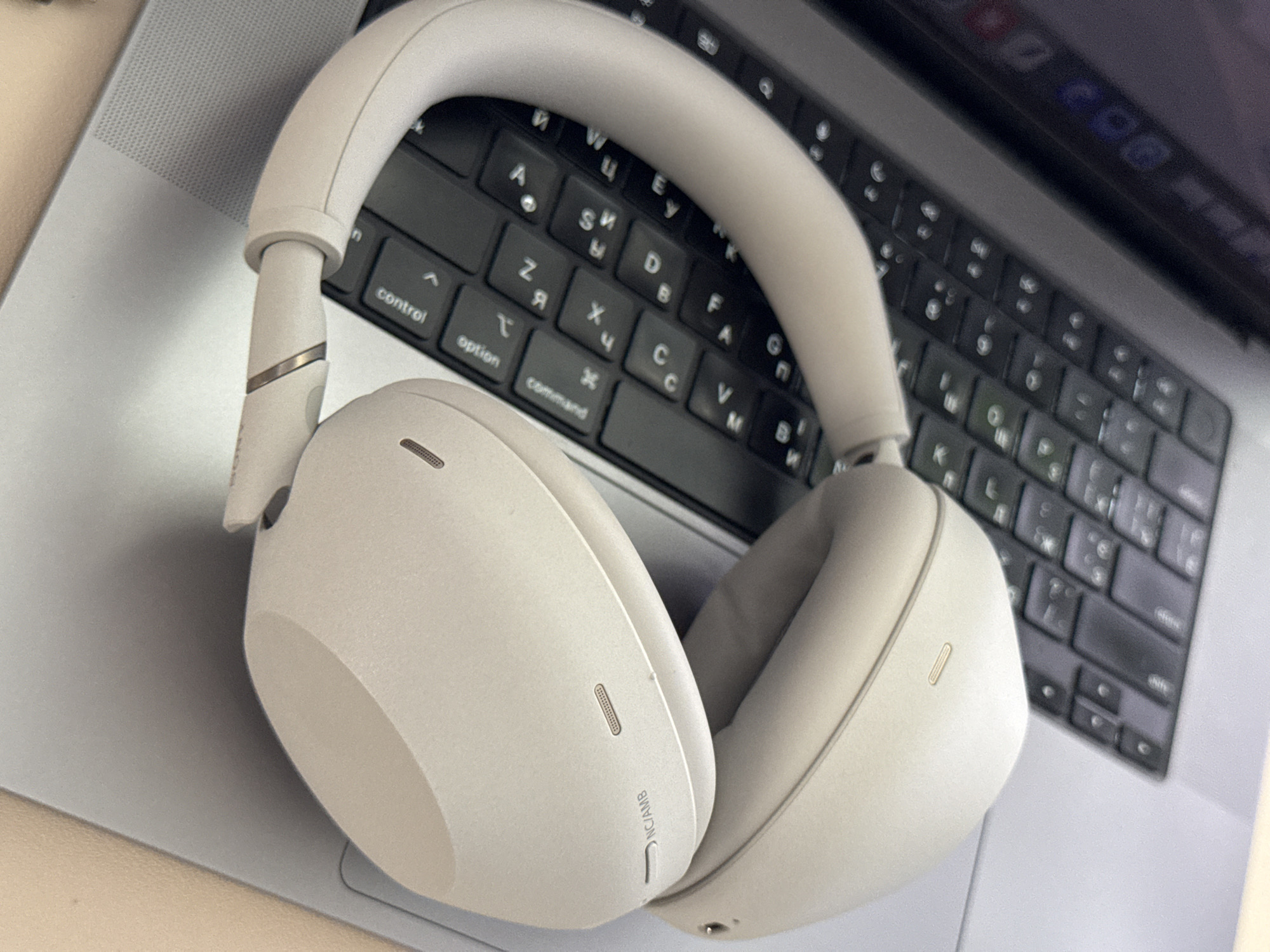 A light-colored pair of over-ear headphones rests on the keyboard of an open laptop. The laptop keyboard has black keys with silver lettering and symbols. The headphones are positioned with one ear cup covering a portion of the keyboard and the other ear cup slightly off to the side. The laptop is silver, and its screen is visible in the background, displaying a dark, blurred image. The background lighting is soft, casting a gentle shadow. The overall scene suggests a work or study environment.