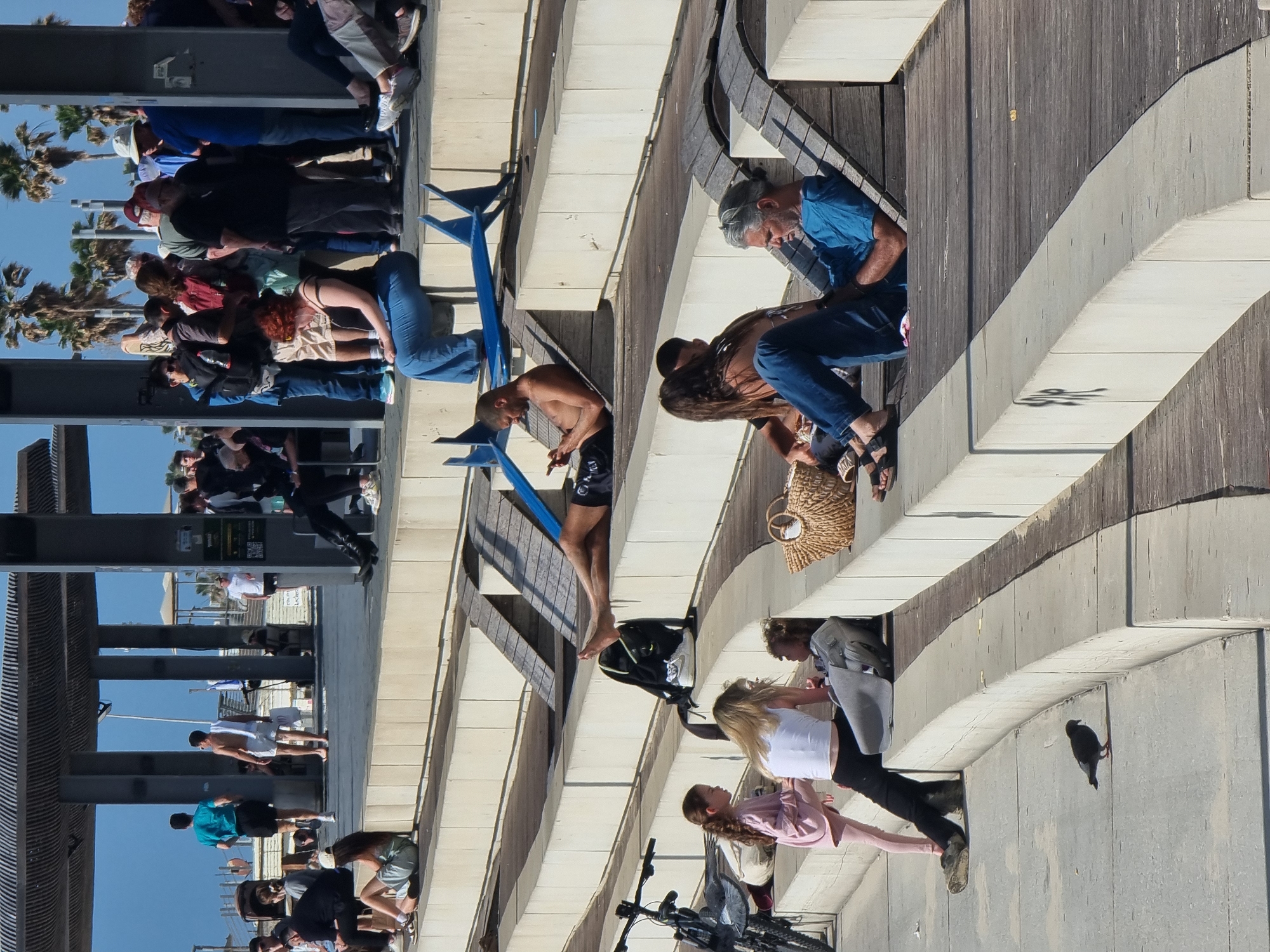 This image captures a vibrant, sunny outdoor scene in Tel-Aviv, Israel, depicting a lively public space, likely a promenade or boardwalk such as the Tel Aviv port (Namal Tel Aviv).
The scene is bustling with numerous people of various ages, dressed in casual summer attire. On the left side, a queue of individuals, including men and women, extends behind blue metal barriers. They appear to be waiting, possibly for an attraction or service. Many wear hats and sunglasses to shield from the bright sun. A shirtless man in black shorts stands near the front of this group, engaged with his phone. Further in the background, more people are visible under large overhead structures, with palm trees dotting the distant horizon, characteristic of a coastal city.
On the right side of the image, the atmosphere is more relaxed. Individuals are seated or resting on multi-tiered wooden and concrete ledges. An older person with grey hair, wearing a blue shirt and jeans, sits, seemingly focused on a device. Nearby, a woman with long dark hair also rests. Further down, a woman in a white top and black pants stands beside a young girl in a pink outfit. Personal items such as a black backpack and a woven basket bag are placed on the ground. A bicycle is visible in the foreground. On a lower concrete ledge, a dark bird, possibly a pigeon, is perched.
The time of day is clearly sunny, with strong, defined shadows indicating midday or early afternoon. The sky is clear, suggesting pleasant weather. Architectural elements consist of light-colored concrete pathways and ledges contrasted with dark wooden planking, creating a modern, multi-level public area. In the background, on a structural pillar, an information sign with a QR code is partially visible. The overall impression is one of a busy but leisurely day in a popular public area.