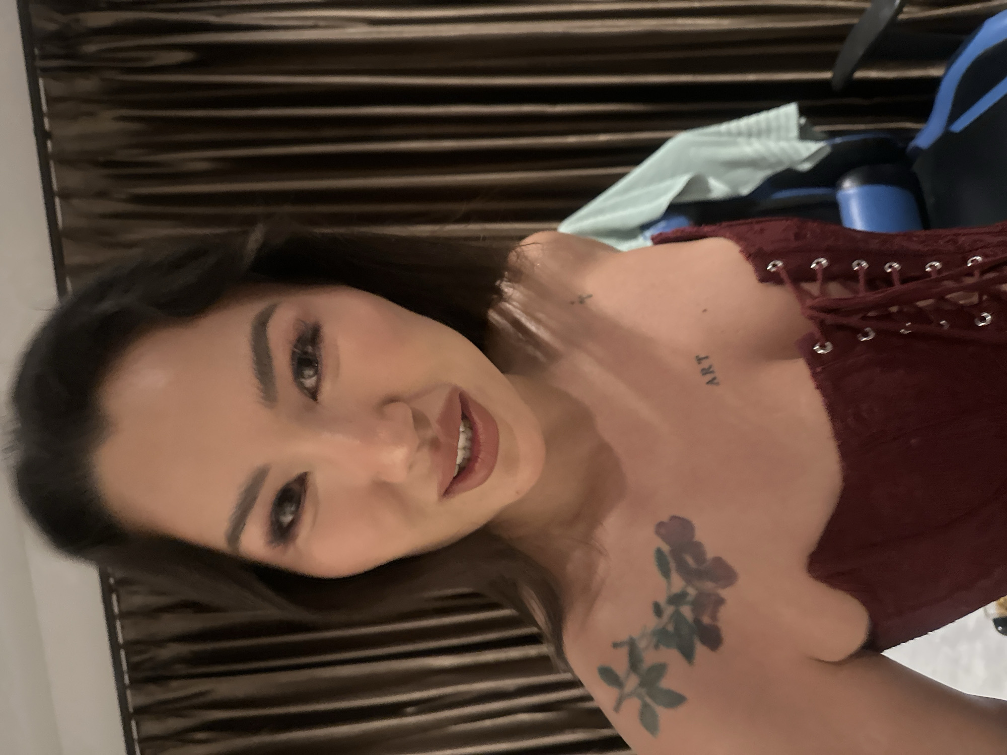 A close-up, medium shot captures a woman's face and upper body against a backdrop of dark brown, vertically striped curtains. The woman, with dark hair and pronounced eye makeup, looks directly at the camera with a slight smile. She is wearing a dark red, lace-up corset. On her left shoulder, a floral tattoo is visible, and on her right collarbone, a small tattoo with the word "ART" is present. Her skin tone is fair. The lighting suggests an indoor setting, possibly a bedroom. The image is taken from a slightly low angle. No specific details about North Kuta, Indonesia, are discernible from the visual information provided.