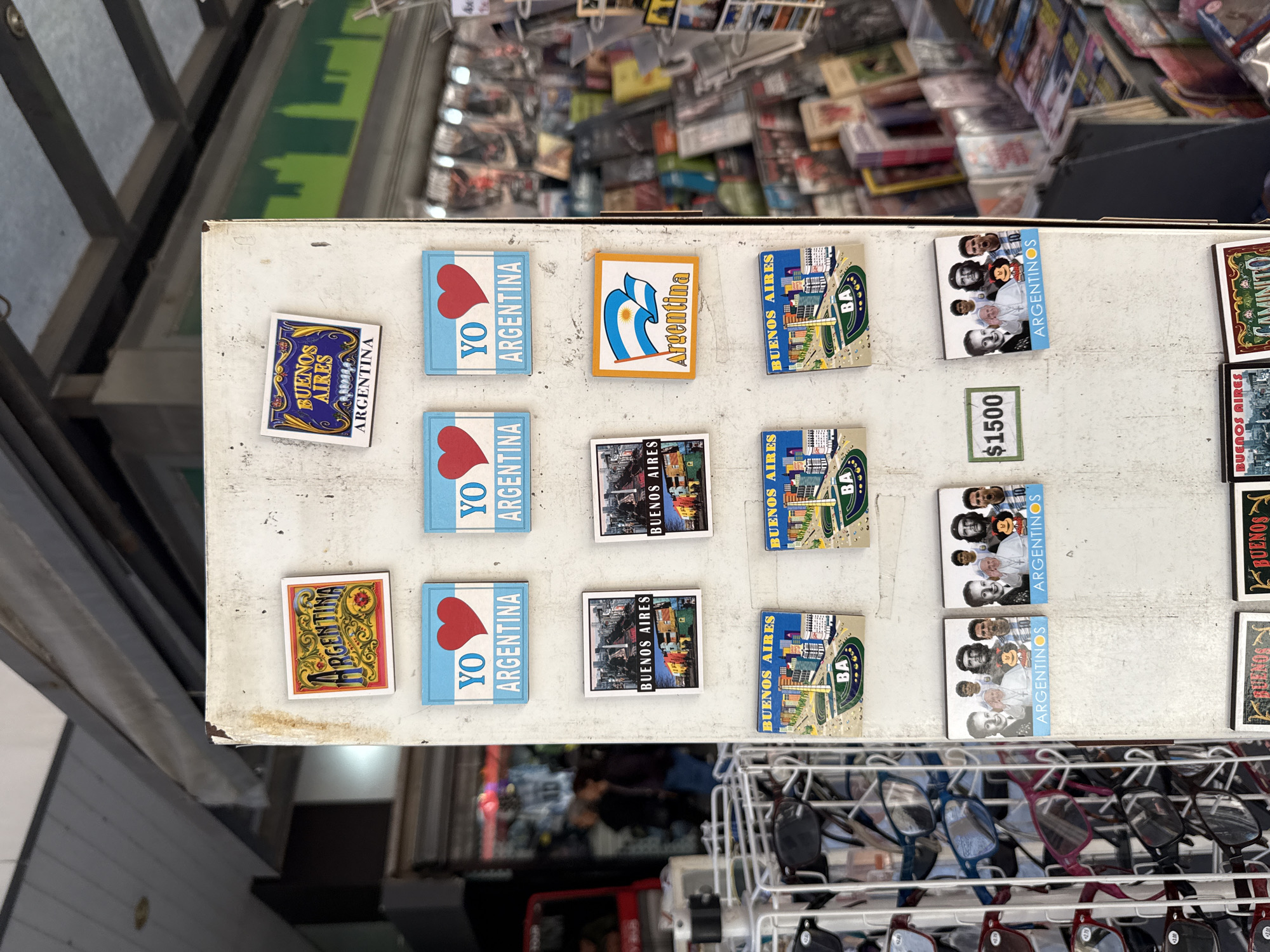 The image shows a collection of souvenir magnets for sale in Buenos Aires, Argentina. The magnets are displayed on a white surface and feature various designs, including the Argentine flag, the words "Buenos Aires" and "Argentina," images of the city skyline, and caricatures of famous Argentinians. Some magnets have prices written on them, such as "$1500." Beneath the magnets, there is a display of sunglasses for sale. In the background, there are more souvenirs and general street scene items. The overall setting suggests a tourist area in Buenos Aires.