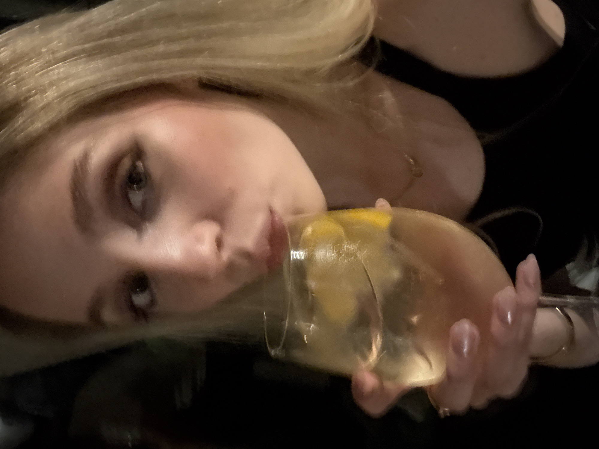 A fair-skinned blonde woman, possibly Caucasian, holds a glass of light-colored liquid, likely a cocktail, to her lips. The drink contains what appears to be a lemon slice. She is wearing a dark-colored top, and jewelry, possibly a gold necklace and ring. The background is dark and indistinct. There is no verifiable evidence to conclusively determine the location of the city of Mexico City, Mexico in the image.