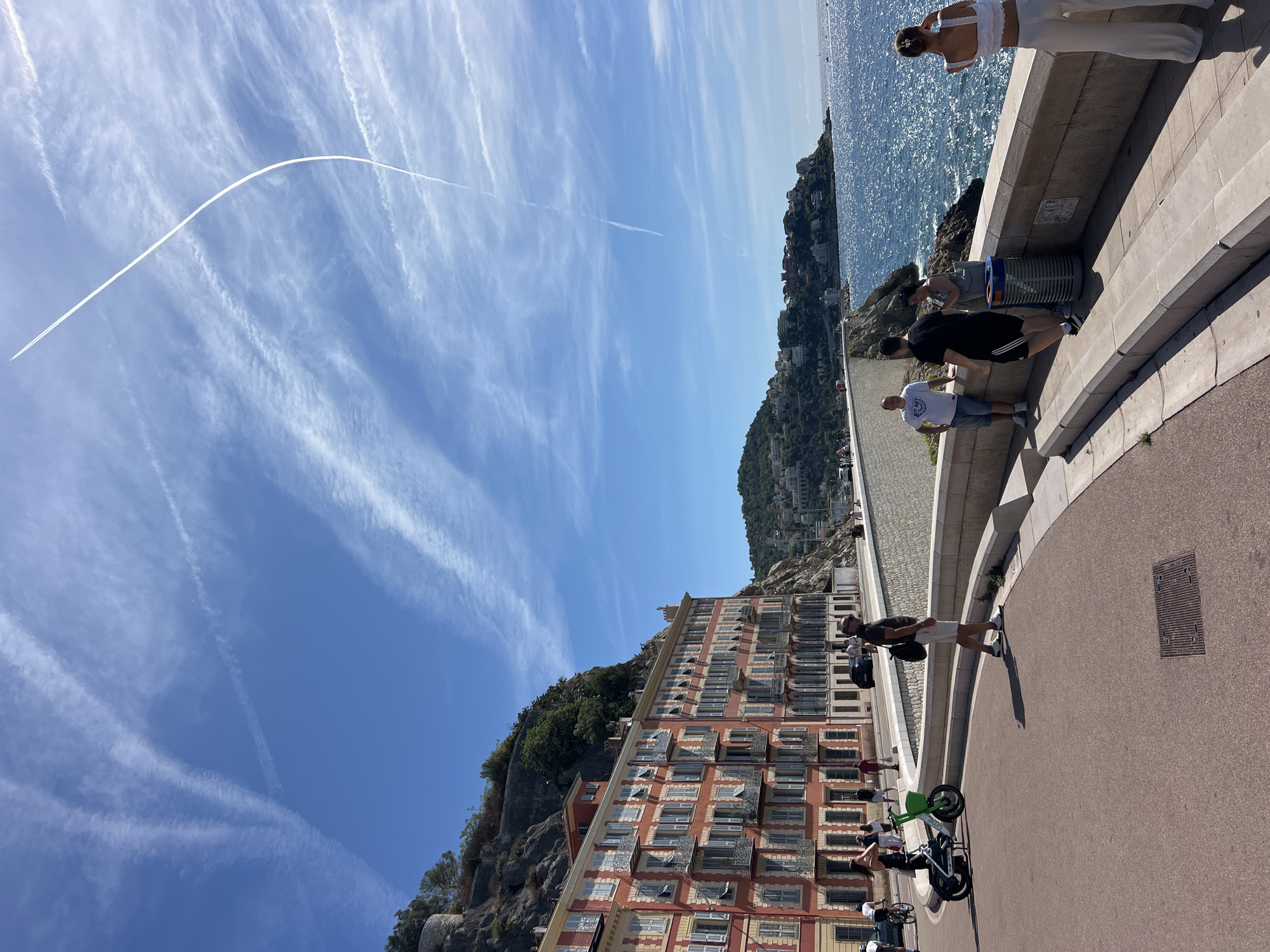 The image depicts a sunny day along the Promenade des Anglais in Nice, France. In the foreground, a reddish-pink, multi-story building with numerous windows and balconies lines the street, with a steep, rocky hillside covered in vegetation rising behind it. A paved walkway with a low stone wall separates the buildings from the sparkling blue Mediterranean Sea on the right. Several people are visible on the walkway. On the left, a woman on a white and green electric scooter is parked. Further along the walkway, a man in a dark t-shirt and white shorts stands with a backpack. To his right, a group of three people are gathered near a trash bin, with two men and one woman. On the far right, a woman in white pants and a white crop top stands facing the sea, with her back to the camera. The sky above is a clear blue, with scattered white clouds and a prominent contrail from an aircraft forming a curved line across the upper portion of the image. The overall scene conveys a sense of leisure and activity in a coastal urban environment.
