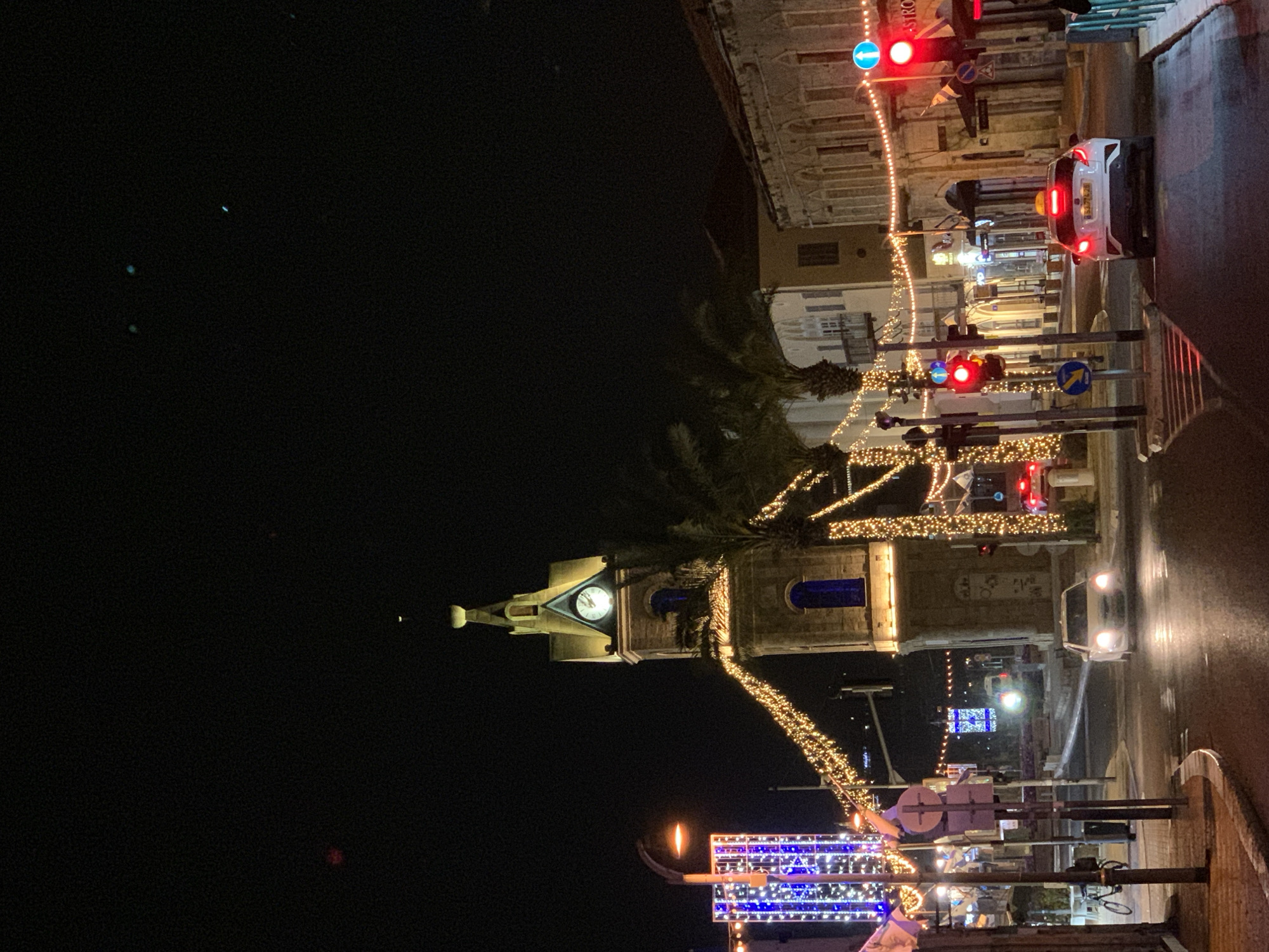 Nighttime scene in Tel Aviv, Israel. A church tower with a clock is illuminated and decorated with lights. Palm trees are adjacent to the church. Streets are also illuminated with lights and street signs. A car is stopped at a red traffic light. Buildings line the street. The sky is dark.