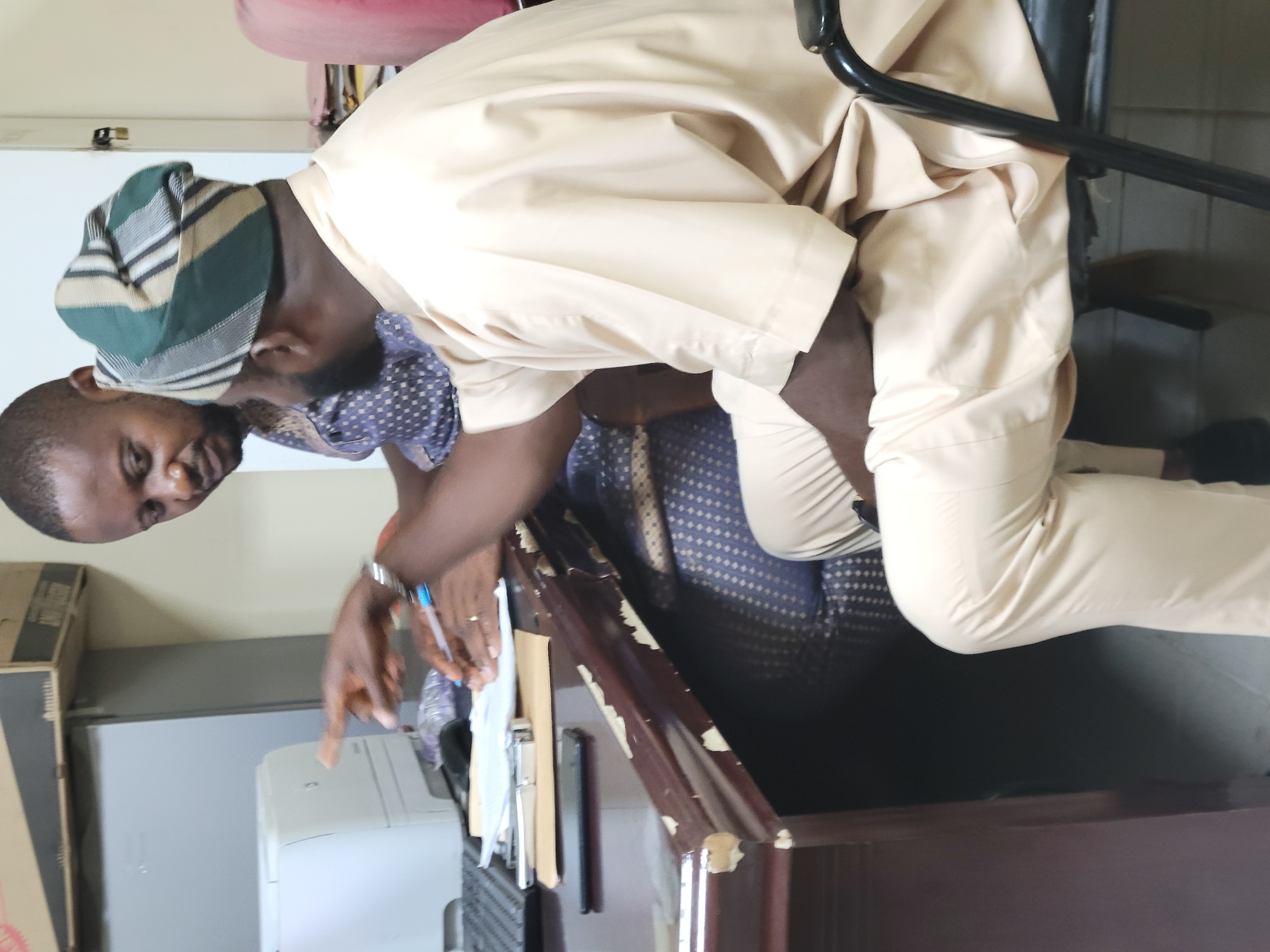 A man is seated at a desk in an office. He is wearing a traditional Yoruba hat called a "fila" and a light-colored traditional attire. He is pointing at something on the desk. A woman sits beside him. There is a printer, documents, and other office supplies on the desk. The location is likely in Abuja, Nigeria, given the subject is wearing a traditional Yoruba attire which is prevalent in Nigeria.