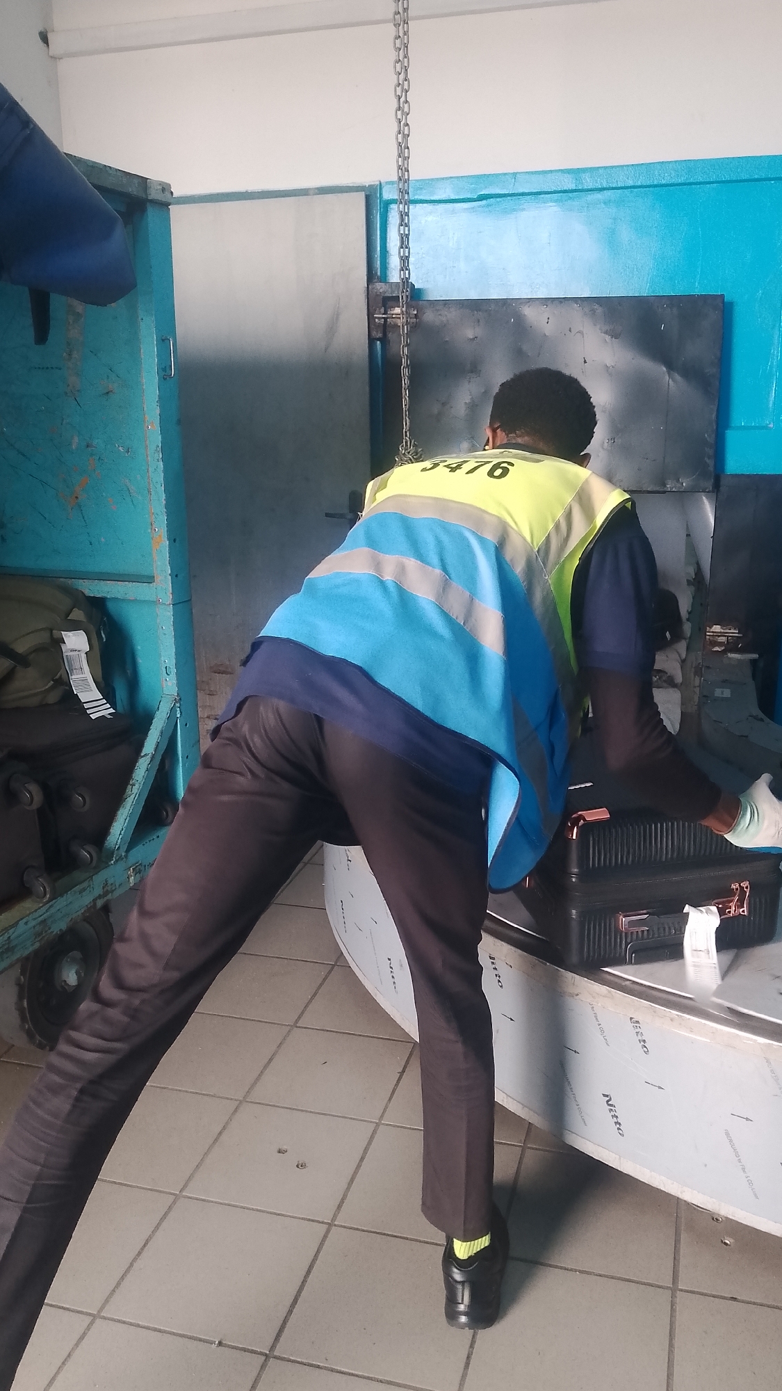 In an indoor setting, likely at an airport or transport hub in Orerokpe, Nigeria, a man wearing a blue short-sleeved shirt, a yellow and blue reflective safety vest with the number "3476" on the back, and dark-colored pants is handling a black briefcase. He is bent over a conveyor belt covered with a silver-colored panel. He is wearing black shoes with bright yellow socks. A metal chain hangs above. In the background are various pieces of machinery and metal containers. The floor is tiled with light gray tiles.