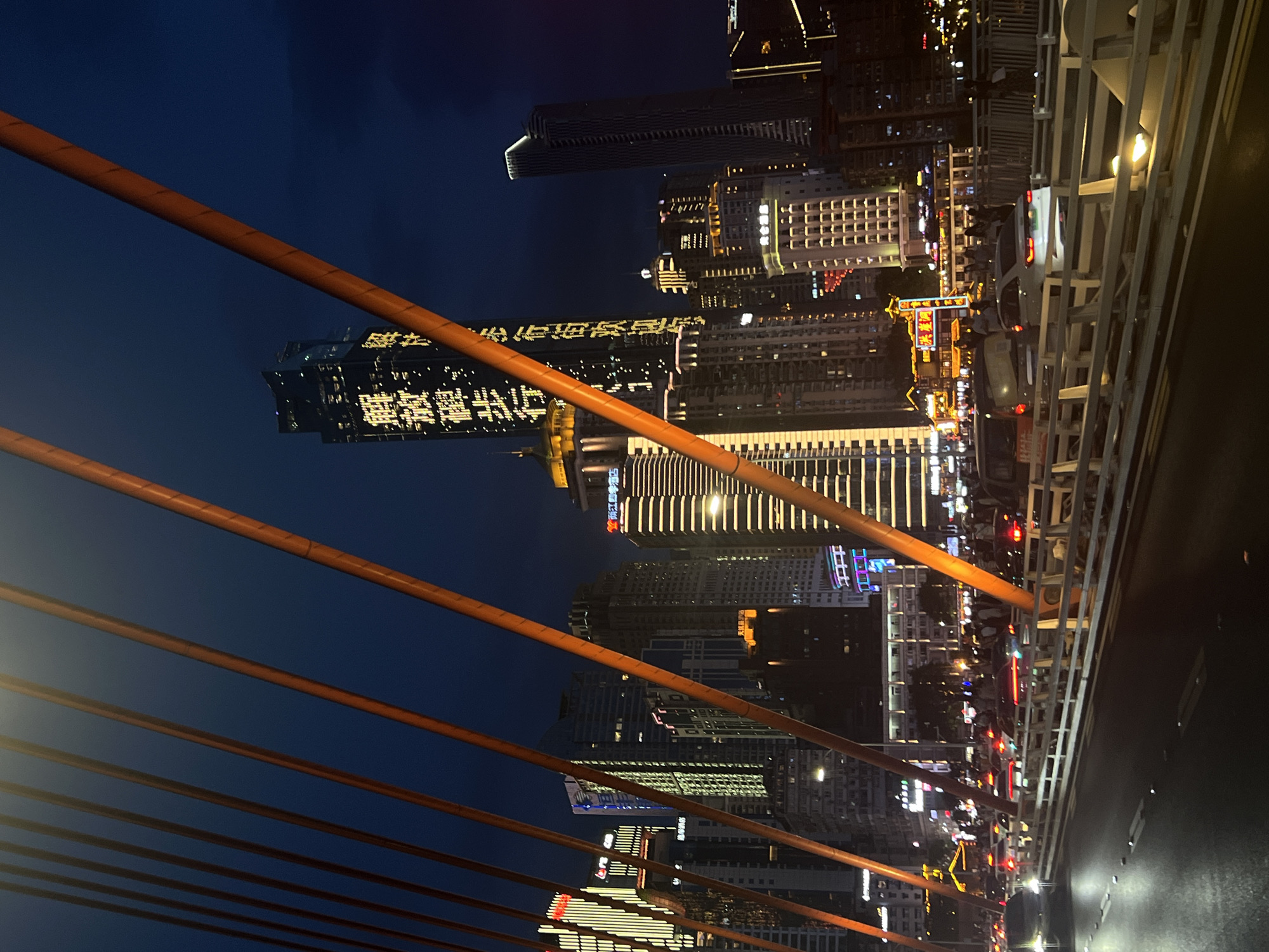The image depicts a nighttime cityscape in Yuzhong District, China, viewed from a bridge. The foreground features a road with several cars in motion, their headlights and taillights illuminated. A white guardrail separates the road from a pedestrian walkway, where a crowd of people is visible. Dominating the background are numerous illuminated skyscrapers of varying heights and architectural styles, showcasing a dense urban environment. Several of the buildings feature prominent signage with Chinese characters. The sky is dark, indicating the late hour. Suspended orange cables, part of the bridge structure, diagonally bisect the frame, creating a sense of depth and perspective.