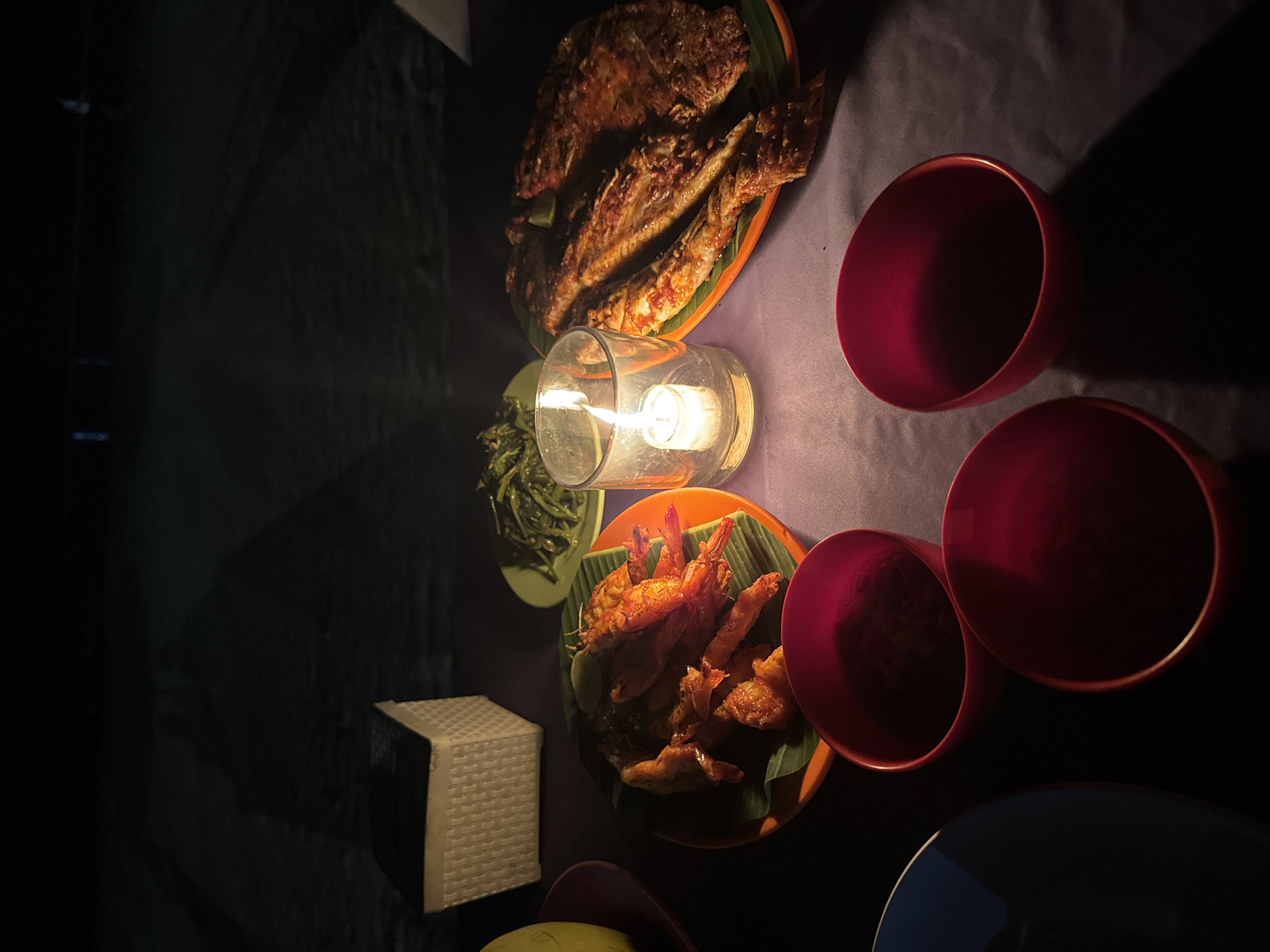 A candle flickers in a glass holder on a table set for a meal at night, likely in Kuta Selatan, Indonesia, given the setting of a beachfront dining experience. The table is covered with a dark purple cloth and illuminated by the candle and ambient light. In the foreground, three red bowls are partially visible, one containing a dark substance. Behind them, a plate of grilled fish, seasoned with red spices, is arranged on banana leaves. To the left of the fish, a plate of fried shrimp, also seasoned and served on banana leaves, is presented. A green leafy vegetable dish is visible in the background, along with a small white woven box. The overall scene suggests an outdoor dining experience at dusk or night, possibly on a beach, with the dark background and shadows indicating the time of day and the surroundings.