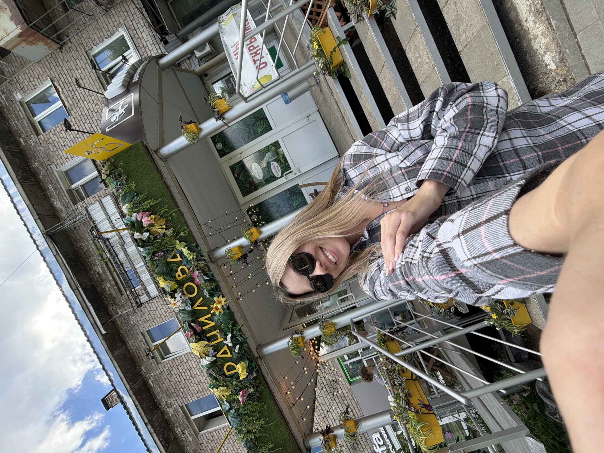 A woman with blond hair and sunglasses sits on a grey metal railing in front of a building. She wears a grey and white plaid shirt. Behind her is the facade of a building with a sign in yellow letters reading "DANKOV". The building also has greenery and floral decorations. The location is the city of Chaikovsky, Russia.