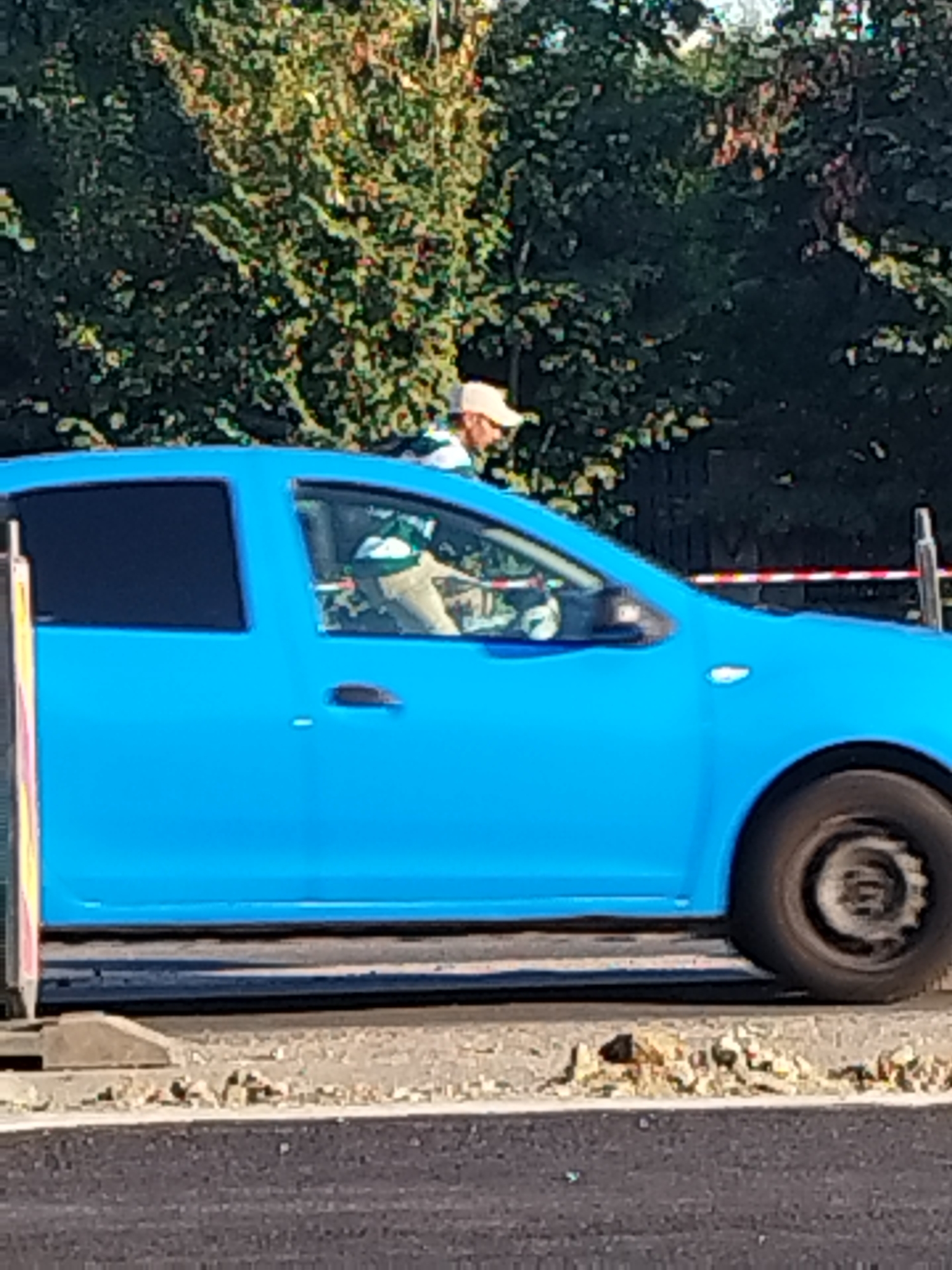 A person wearing a white baseball cap and a striped green and white shirt is driving a blue car. The car is on a paved road with some construction debris to the right. In the background, there are trees. The image appears to have been taken in Bucharest, Romania.