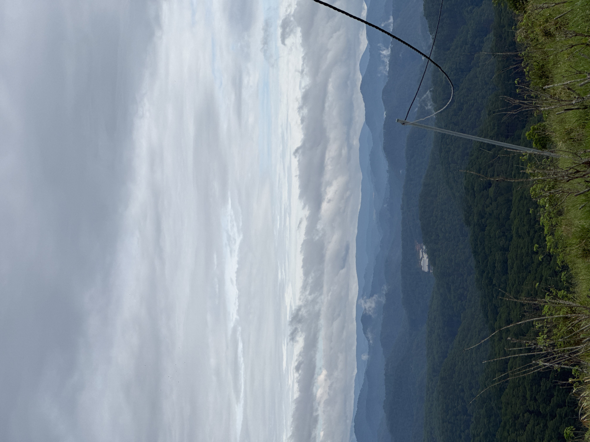 From a high vantage point in Trường Thọ, Vietnam, the image captures a landscape featuring a cloudy sky, layered mountains, and lush vegetation. Cables, possibly related to a cable car or zipline, cut across the scene. The sky is predominantly overcast, with varying shades of gray. The mountains recede into the distance, displaying different hues of blue and green due to atmospheric perspective. The foreground shows verdant plant life on the edge of the overlook. A clearing with lighter structures is visible in the middle distance.
