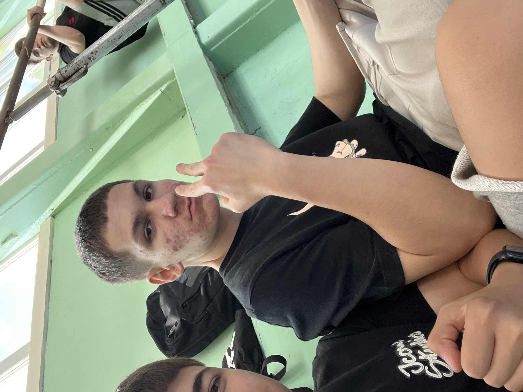 The image shows a medium shot of a young man with short brown hair. He is wearing a black t-shirt and giving the peace sign with his right hand, which is held close to his face. He appears to be sitting next to another person. A partial view of a person in a black and white Adidas tracksuit is visible in the background on a window ledge. The background consists of a pale green wall. The location is potentially in Asenovgrad, Bulgaria.