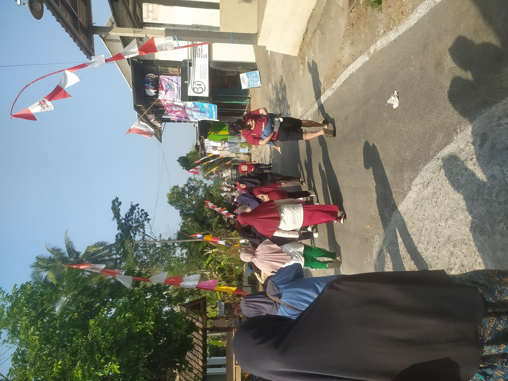 In Lopait, Indonesia, a group of people are walking down a street adorned with red and white flags, likely celebrating a national holiday. The street is paved and has a white line marking the edge. Buildings line the right side of the street, some appearing to be shops or residences. A man in a red shirt is carrying a child on his shoulders. Most of the people are wearing casual clothing, and some women are wearing hijabs. The sky is clear and blue, and the sun is casting long shadows on the street.