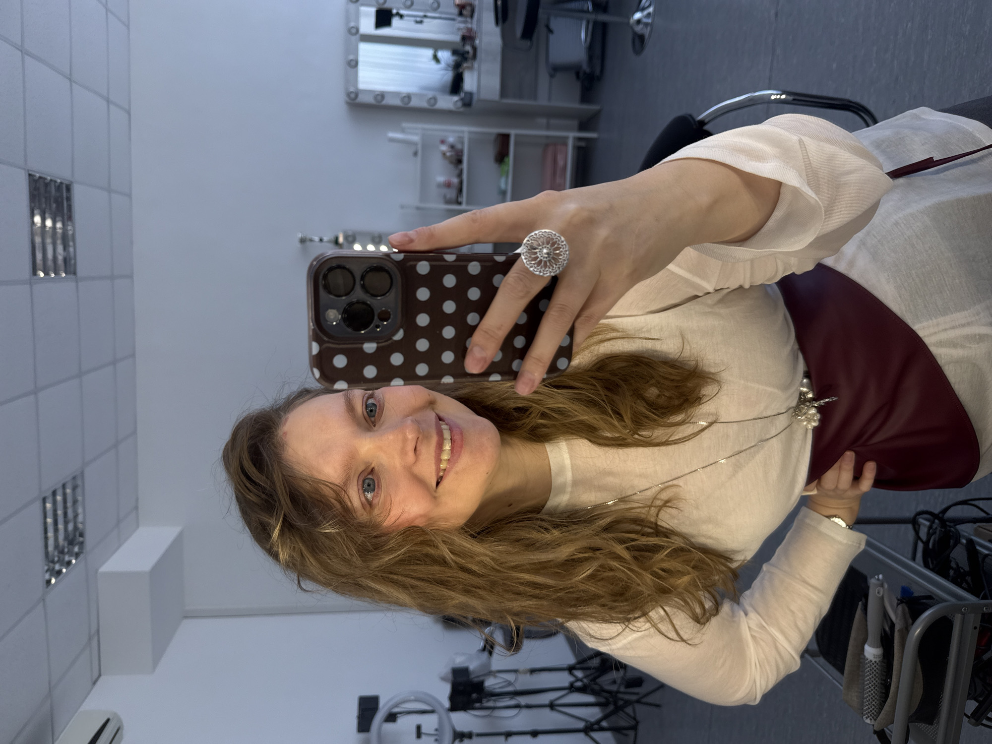A woman is taking a selfie in a room with white tile walls. The woman has blonde hair and blue eyes. She is wearing a white shirt and a large silver ring. She is holding a smartphone with a brown and white polka dot case. The room contains a makeup mirror with lights, a shelf, a salon chair, and a rolling cart with styling tools. There is also a Ring light on the floor. I have no information to verify the location is Izhevsk, Russia.