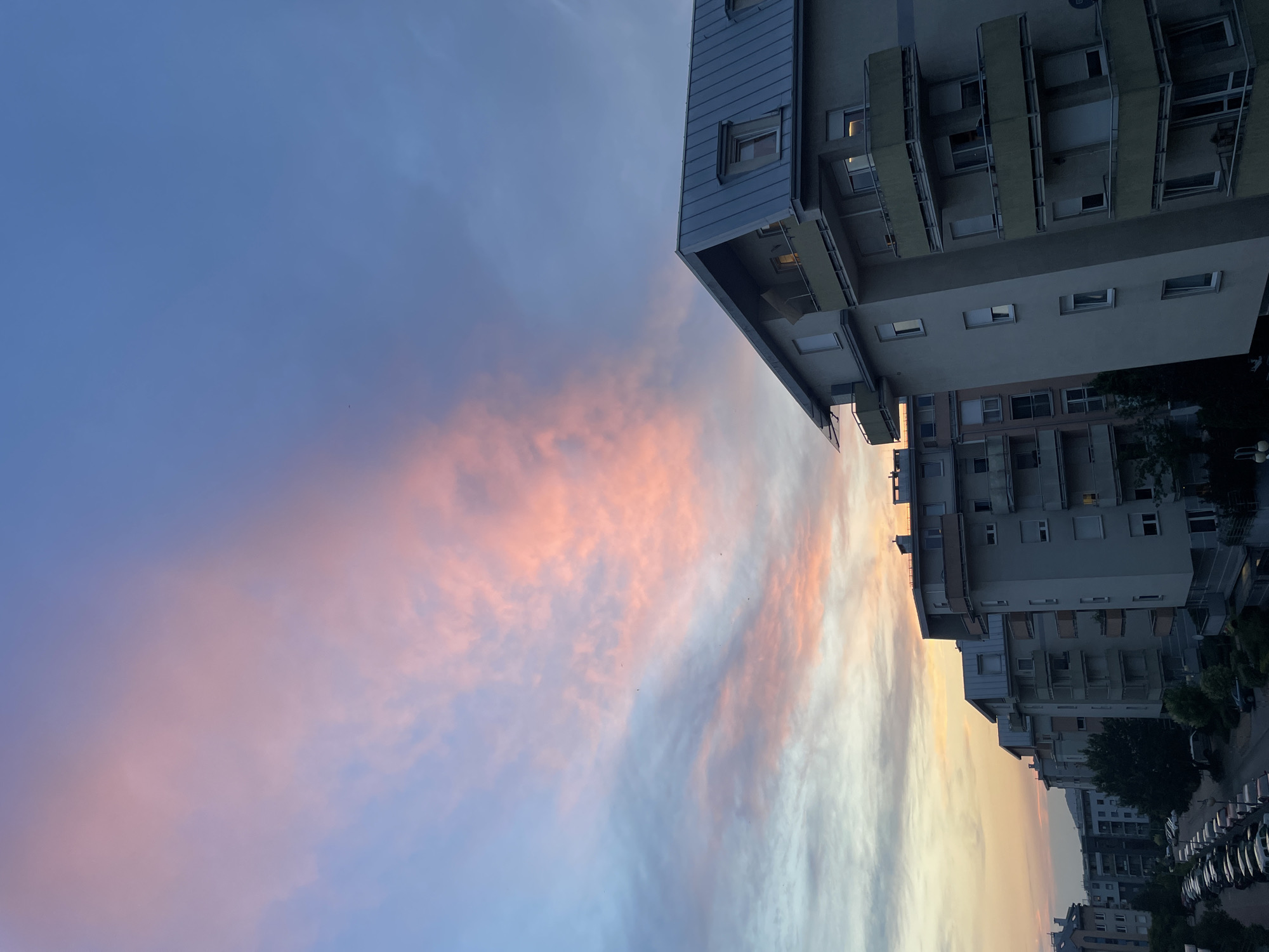 The image captures a street-level view of apartment buildings against a twilight sky. The sky is a gradient of blues and purples, with streaks of soft pink and orange clouds indicating either sunrise or sunset. The buildings are modern, with multiple stories, balconies, and many windows, some of which are illuminated, suggesting that people are home. The architecture is functional and uniform, with a repeating pattern of balconies and windows.
In the foreground, a line of parked cars is visible, adding to the urban setting. Trees are also interspersed between the buildings, their leaves dark against the fading light. The overall scene evokes a sense of calm and domesticity, typical of a residential area at dusk. The orientation of the image is such that the buildings appear to rise from the bottom right of the frame, with the sky occupying the majority of the left side. There is no visible text in the image. The time of day is clearly either dawn or dusk due to the colorful sky and the presence of artificial lights within the buildings. The weather appears clear, with the clouds being a feature of the atmospheric light display. The location is a dense urban environment, likely a residential neighborhood.
