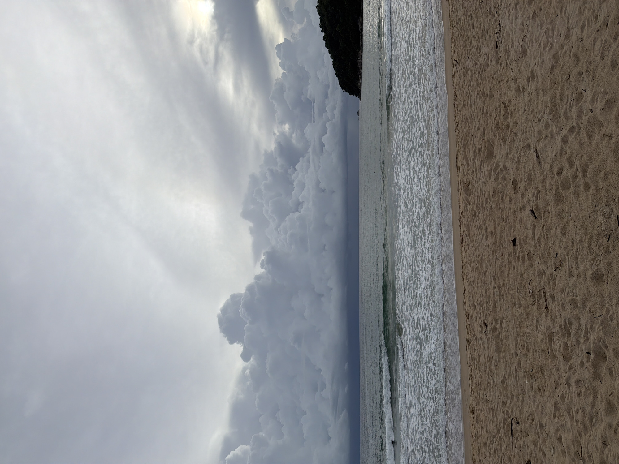 The image depicts a beach scene with a dramatic sky. The foreground is a sandy beach with footprints and some dried plant debris. To the left of the sand is the ocean, with gentle waves rolling in. The water appears calm and shallow near the shore, with a foamy white line marking the breaking waves.
Beyond the ocean, a dark, tree-covered hill or cliff juts out into the scene from the right side, creating a distinct line between the sea and the sky. The sky itself is the most striking element, filled with large, turbulent, grey clouds. Sunlight breaks through in some areas, creating bright, almost white patches, suggesting either a sunrise or sunset, or a break in an overcast day. The overall mood is dramatic and somewhat moody due to the heavy clouds, but the presence of the beach and ocean suggests a natural, perhaps serene, setting.
There are no visible people or objects on the beach or in the water. The image appears to be taken during the daytime, likely late afternoon or early morning given the light and cloud formation. The location is identified as Choeng Thale, Thailand. There is no visible text in the image.