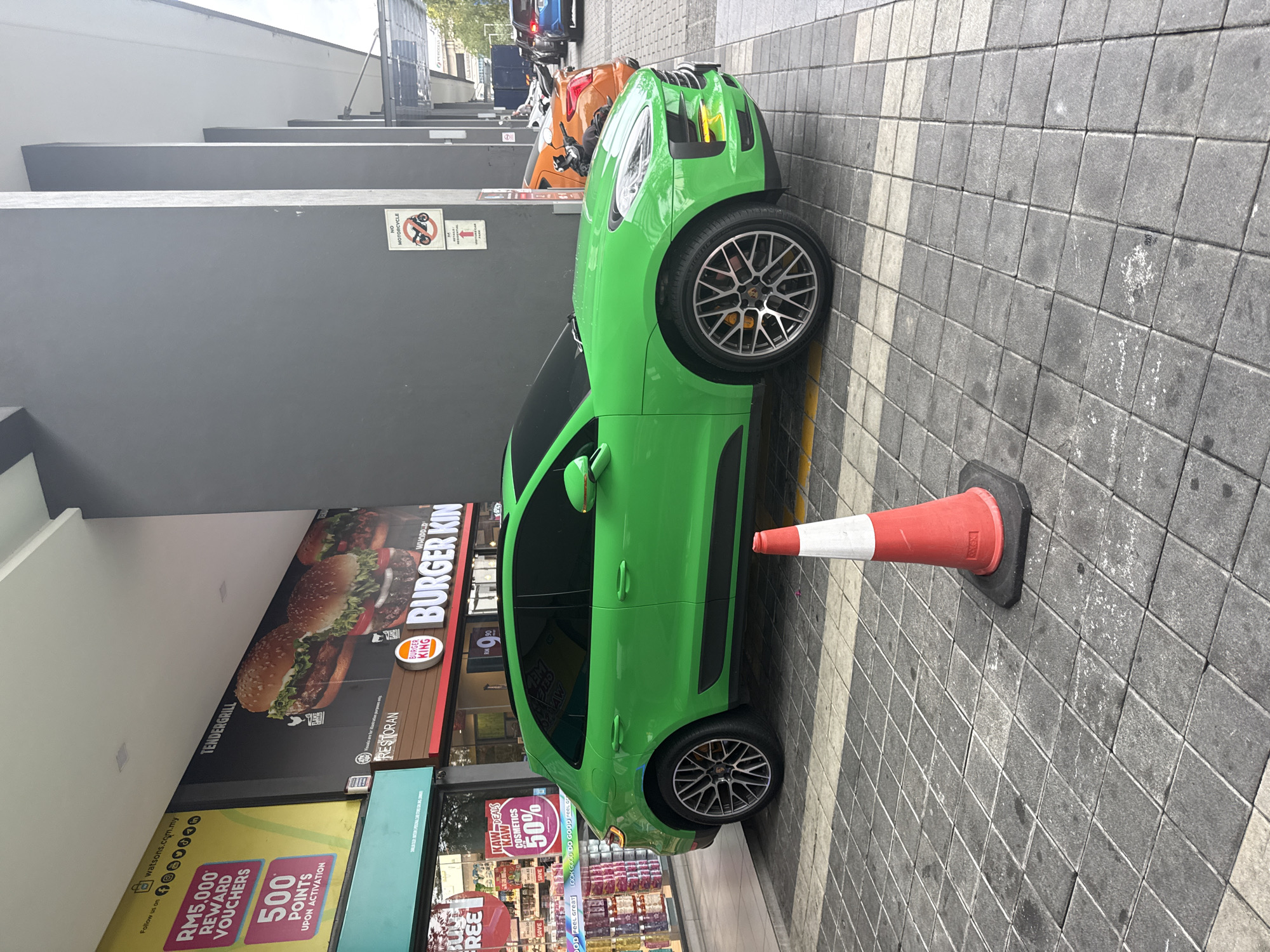 A bright green Porsche Macan is parked along the sidewalk next to a Burger King restaurant and a Watson's pharmacy. The location is in Kuala Lumpur, Malaysia, as indicated by the "watsons.com.my" text on a yellow sign promoting reward vouchers. The sidewalk is paved with gray bricks, and a red and white traffic cone is placed next to the car. The restaurant sign above the car advertises "Tendergrill" burgers. Several motorcycles are parked ahead of the car.