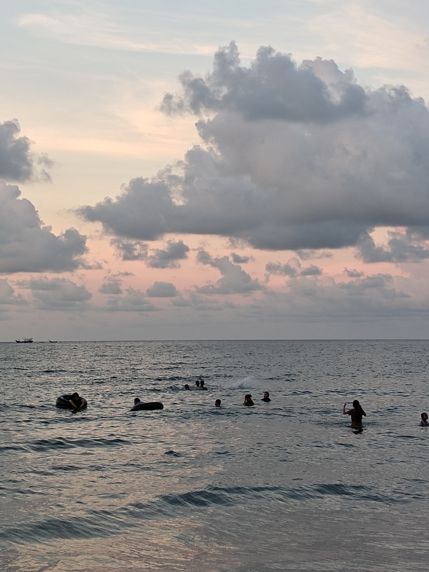 The image shows a seascape with several people swimming in the water. In the distance, there are two small boats. The sky is filled with clouds tinged with pink from the setting sun. The location is Lhokseumawe, Indonesia. Some people are in inner tubes and others are standing or swimming.