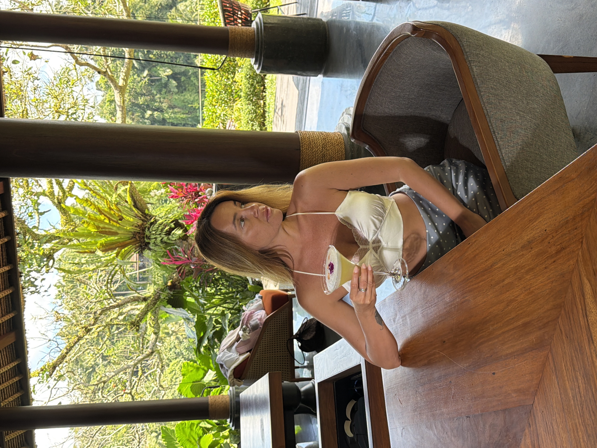 A woman with blonde hair is seated in a gray armchair at a wooden table, holding a cocktail. She is wearing a white satin top and a gray polka-dot skirt. Behind her, lush green foliage and trees are visible through an open-air structure with wooden columns. The location appears to be a resort or restaurant in a tropical setting.