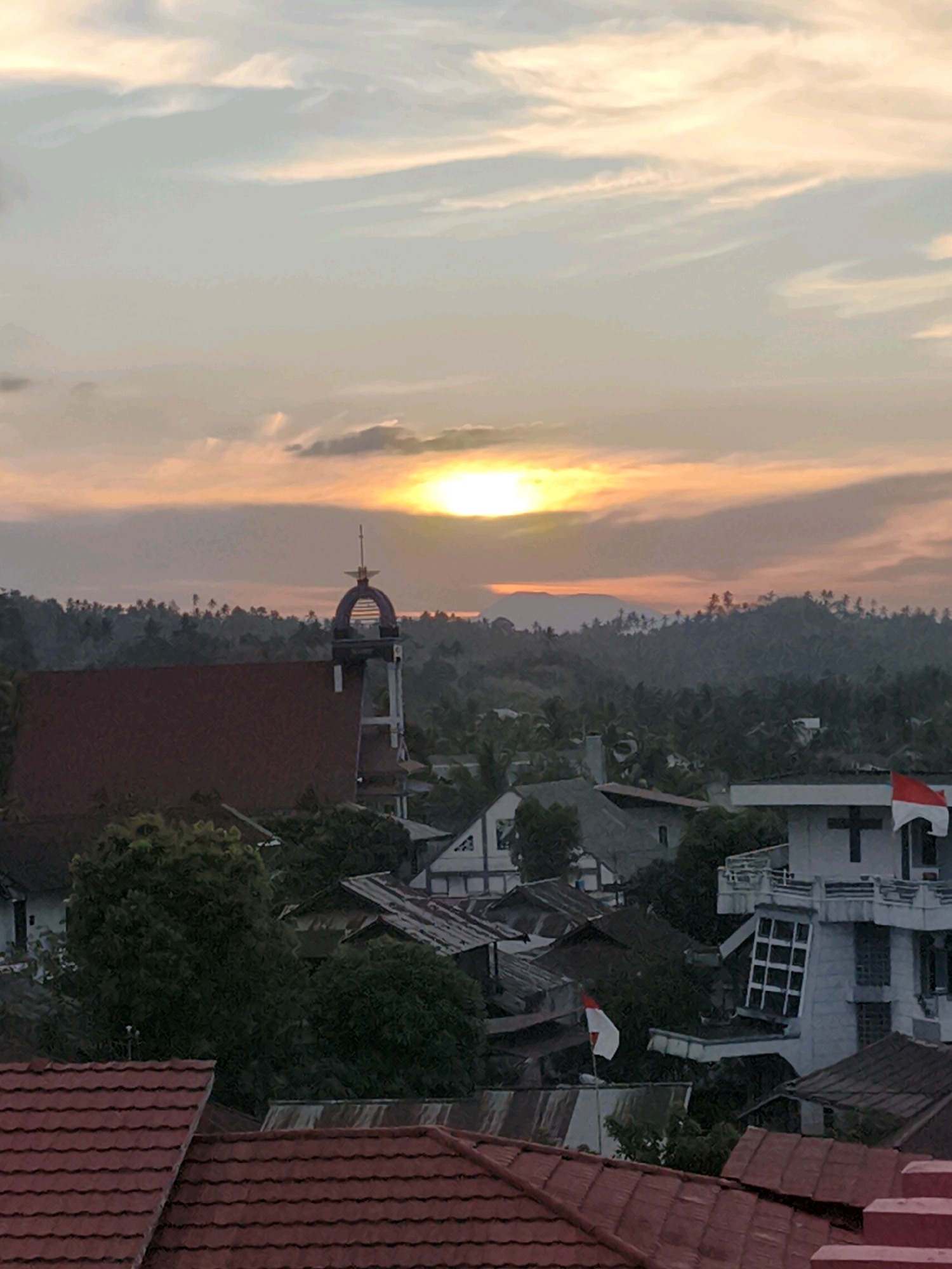 This image shows a sunset over the city of Tombatu Dua, Indonesia. The sky is a mix of orange, pink, and yellow, with some clouds. In the foreground, there are several houses with red tile roofs. In the middle ground, there is a church with a tall steeple. In the background, there are rolling hills covered in trees. A flag, presumably the Indonesian flag due to its red and white colors, is visible on a building. The scene appears to be a peaceful residential area.