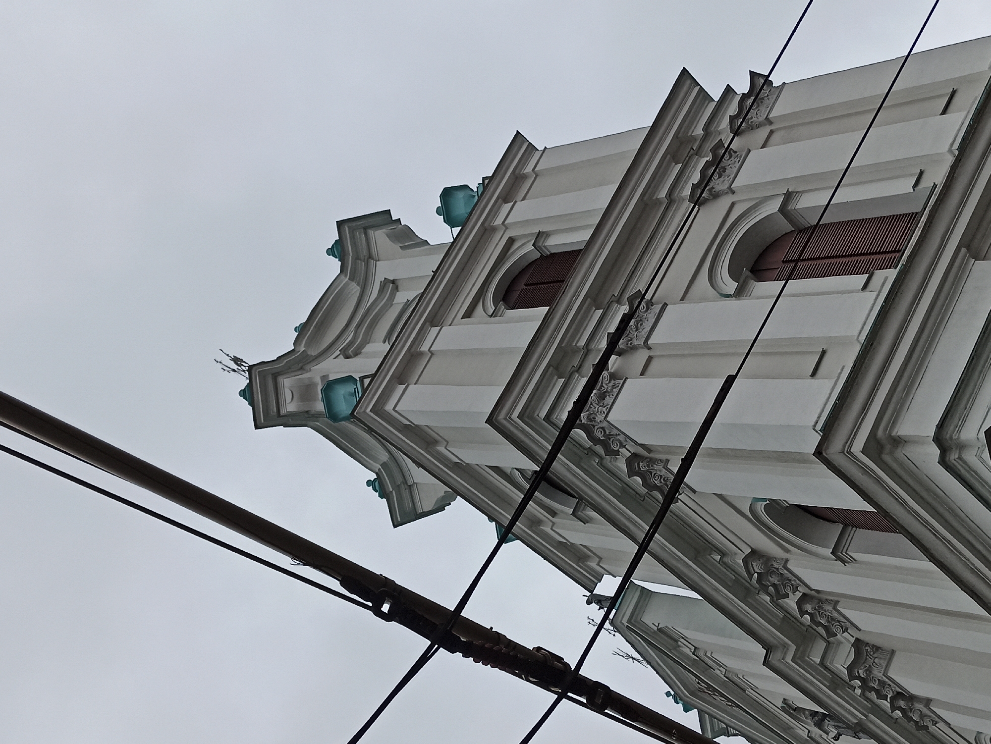 The image shows an upward view of the Church of St. Francis Xavier in Hrodna, Belarus. The architecture features baroque elements, including decorative moldings, curved shapes, and teal domes. The church is painted white, and there are window openings with brown shutters. Electrical wires intersect the view, contrasting with the historical architecture. The sky is overcast.