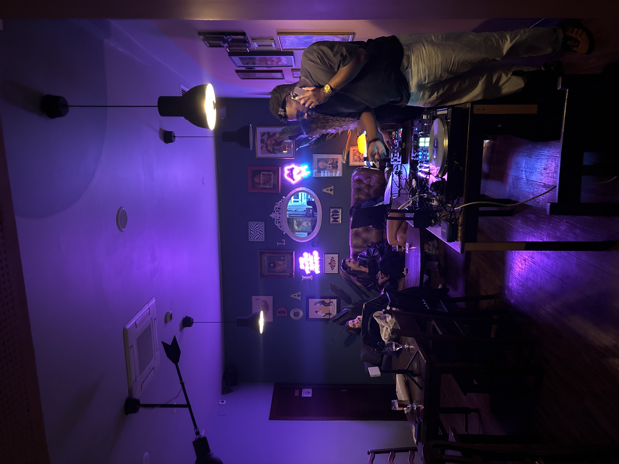 The image shows the interior of a bar or club, likely in Lisbon, Portugal, given the presence of Portuguese writing on the wall. A man is standing behind a DJ setup, using a phone while also operating the equipment. There are several people seated at tables, presumably patrons. The lighting is predominantly purple and dim, with a modern, artistic decor. Artworks and neon signs decorate the wall. A ventilation unit is visible on the wall.