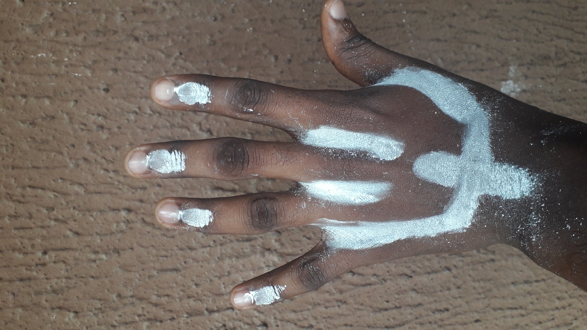 A close-up shows a dark-skinned human hand with white paint or powder markings on the fingers and back of the hand. The fingertips have small dabs of the white substance. Three parallel lines extend across the back of the hand from the index, middle, and ring fingers. A curved shape is painted on the back of the hand near the thumb. The background is a textured brown surface. The image has been taken in Nkpor, Nigeria.