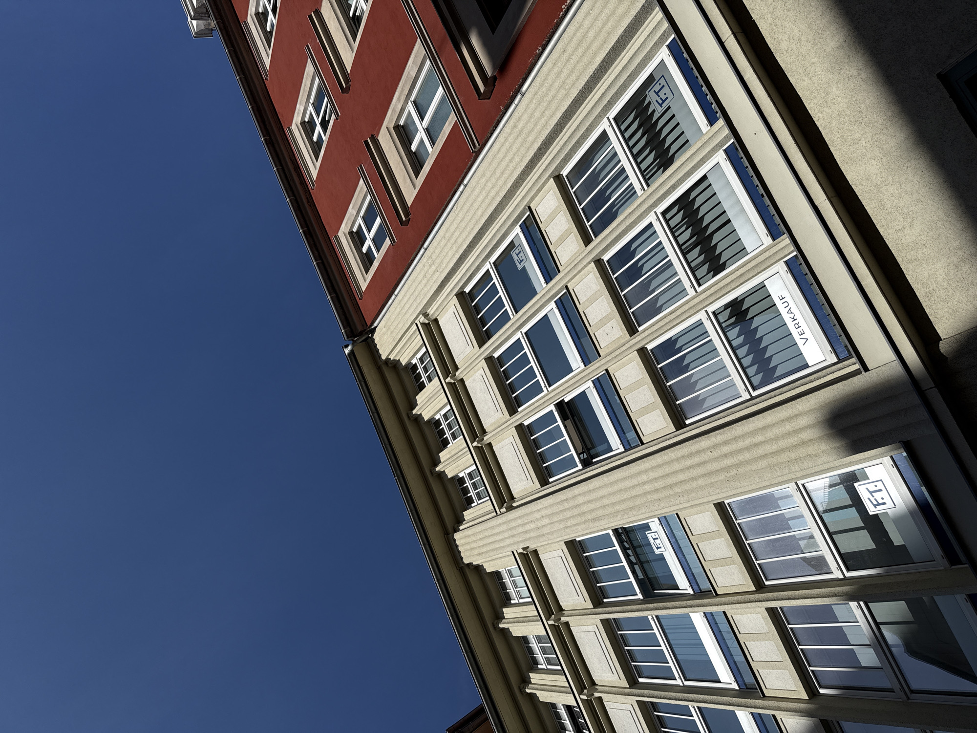 This image captures a low-angle view of two adjacent buildings under a clear blue sky, likely in Görlitz, Germany, given the architectural style. The building on the left features a light-colored facade with a distinct vertical ribbed texture and a grid of numerous windows. The building on the right is painted a vibrant red and also has a series of windows. The angle of the shot emphasizes the upward perspective, with the buildings appearing to recede into the sky. Shadows from the buildings are cast across the facades, indicating a bright sunny day. One window on the left building displays the word "VERKAUF," which translates to "FOR SALE" in German, and a logo with the letters "F.T." within a square. Another window to its right also displays the "F.T." logo. The overall scene is urban and architectural, highlighting the geometric lines and colors of the buildings.
